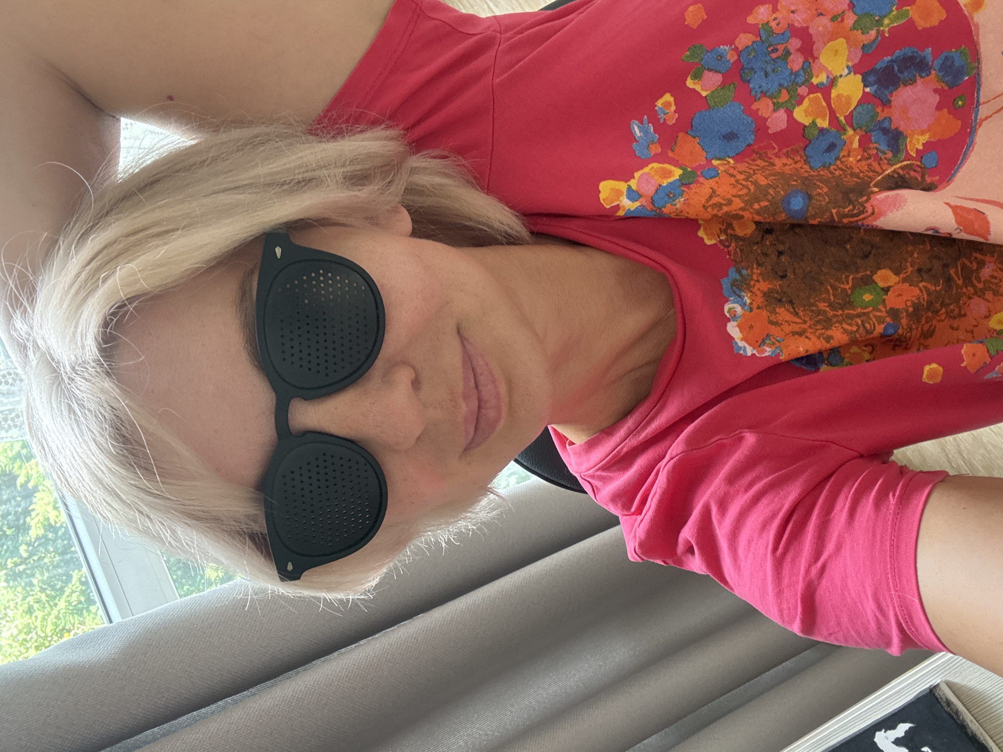 A woman with short, blonde hair wears black, pinhole glasses and a bright pink t-shirt with a floral print. She is indoors, with a window and curtains visible in the background. The lighting suggests it is daytime.
