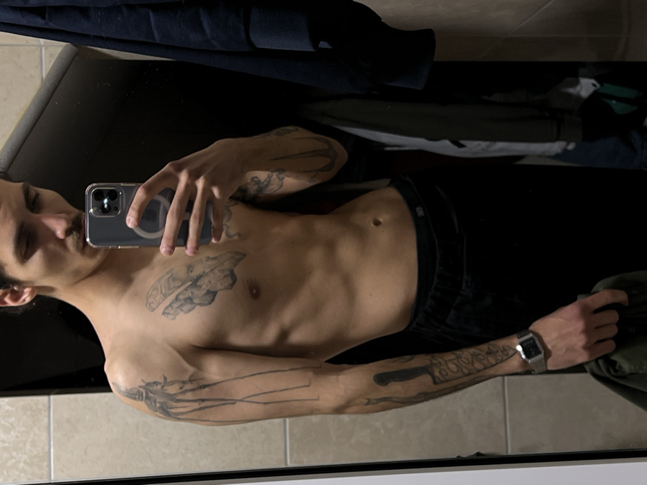 A male individual is depicted from the waist up, captured in a mirror selfie within an indoor setting. The subject, appearing to be an adult male with a lean physique, dark hair, and a mustache, is shirtless. Visible tattoos include a large, stylized, elongated figure on his upper left arm, abstract or fragmented designs on his chest, a kitchen knife or cleaver design on his lower left forearm, and various abstract designs on his right arm. He is wearing dark-colored sweatpants or lounge pants, and a silver-toned digital watch is present on his left wrist. His right hand holds a smartphone with a clear case and visible MagSafe ring, partially obscuring the lower half of his face as he takes the photograph. His left hand is holding a piece of dark green fabric. The environment reflected in the mirror consists of light-colored, possibly beige, square wall tiles on the left and upper frame, and a dark background on the right side which appears to show hanging dark-colored clothing, indicative of a closet or similar storage area within a private residence. The location in the city of Budva, Montenegro, cannot be determined from the visual evidence presented in the image.