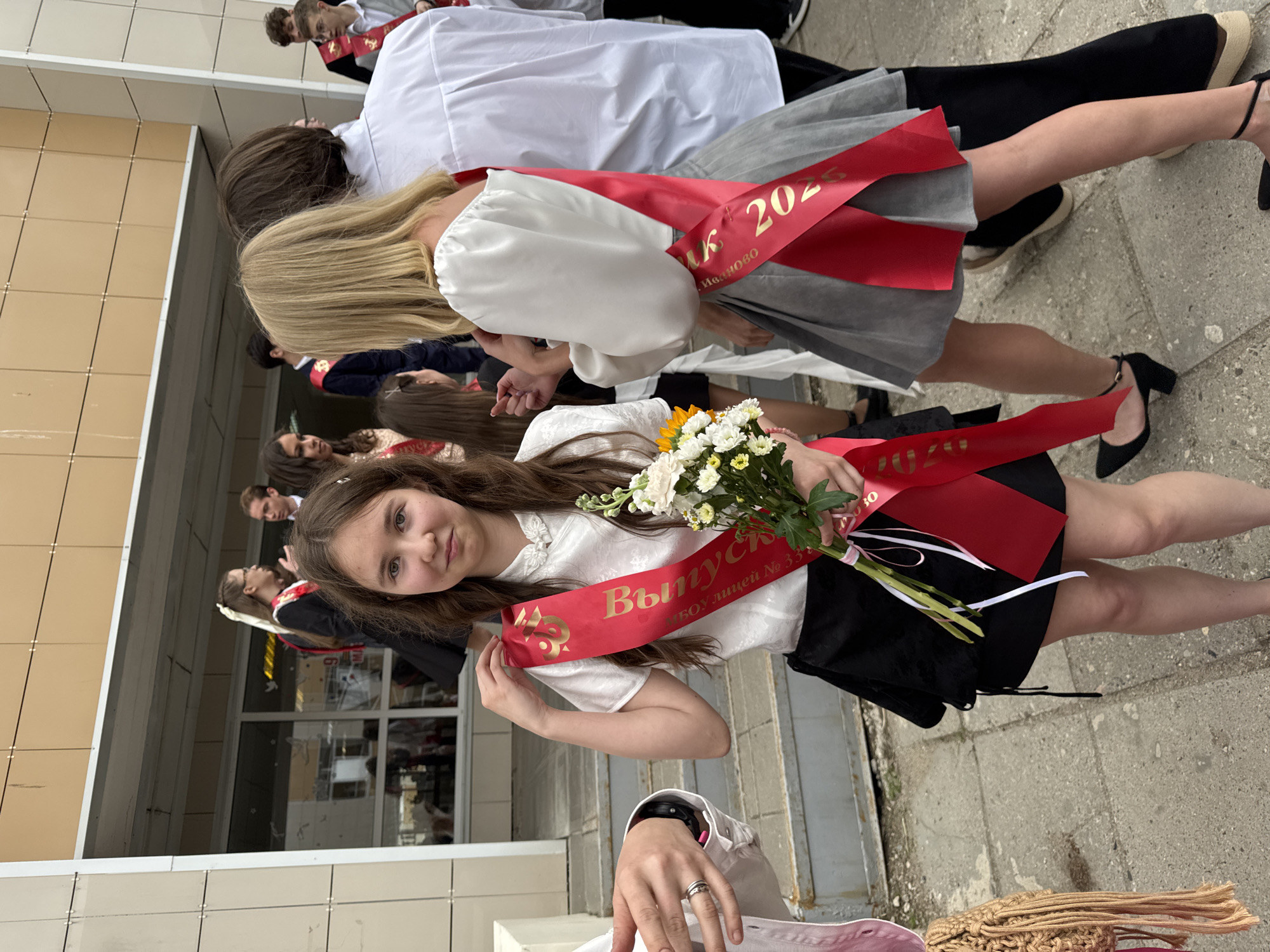 The image depicts a group of graduating students in the city of Ivanovo, Russia. The primary subject is a young girl with brown hair, wearing a white blouse and a black skirt. She is holding a bouquet of white and yellow flowers and is adorned with a red sash that reads "Выпуск 2023" (Graduation 2023) and "МОУ Лицей № 33" (Municipal Educational Institution Lyceum No. 33). Other students are partially visible in the background, wearing similar graduation sashes and outfits. The setting appears to be outdoors, near a building with a tiled facade. The environment suggests a school or educational institution.