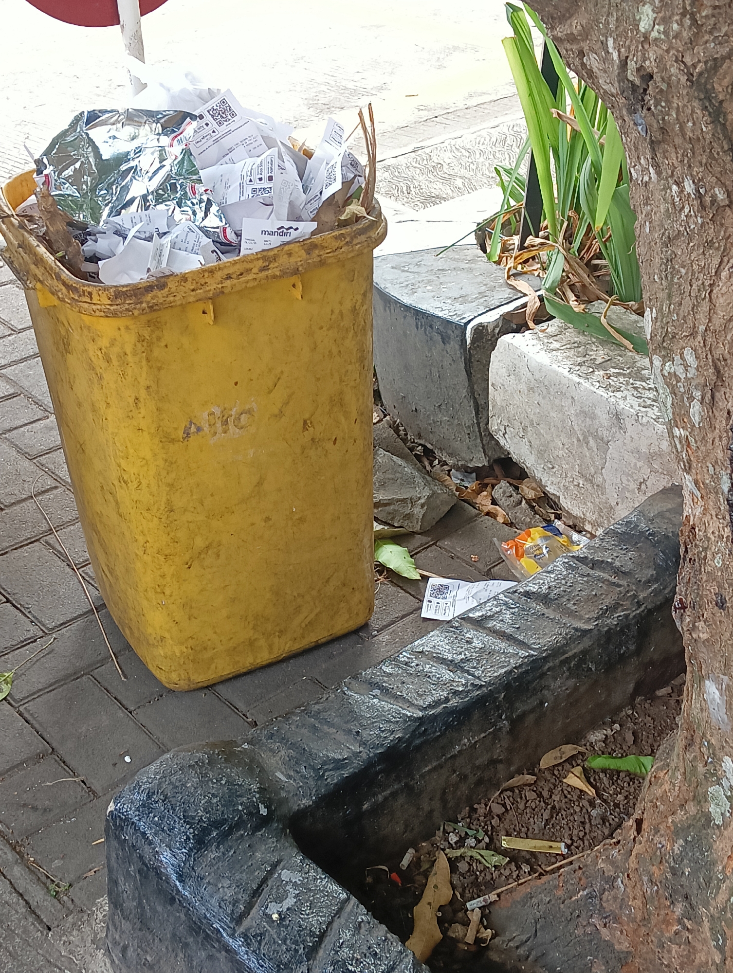 A yellow plastic trash bin, filled with crumpled paper and foil, sits on a brick sidewalk in Cigintung, Indonesia. The bin has "ALIC" written on its side in faded blue marker. Beside the bin are two concrete blocks, one with plants growing around it. A large tree trunk is to the right of the scene. On the ground near the bin, scattered leaves, a QR code receipt, and a small plastic bag with colorful wrappers are visible.