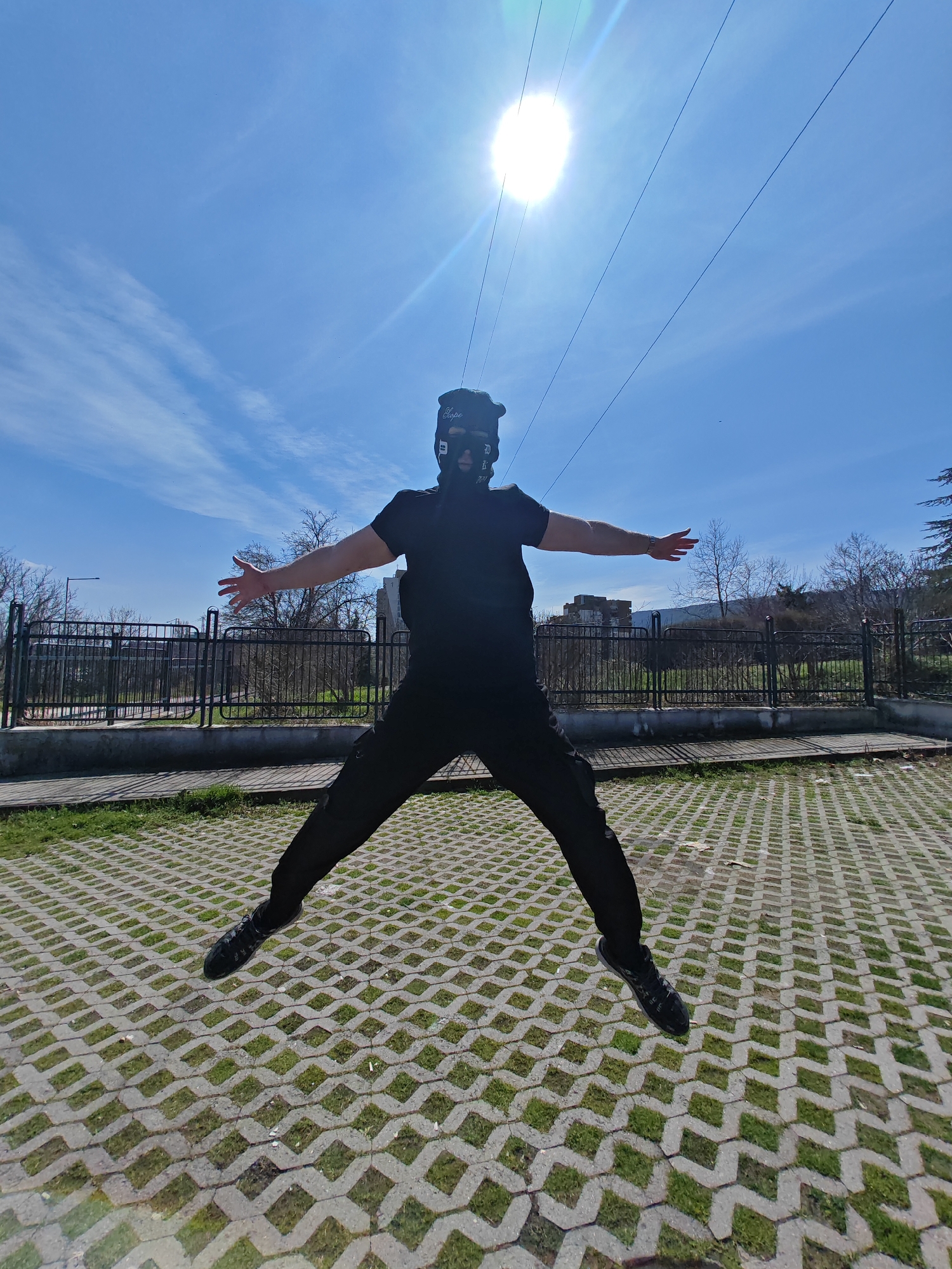 A single individual is captured mid-jump outdoors on a clear, sunny day. The subject is centered in the frame, dressed entirely in black, consisting of a short-sleeved t-shirt, trousers, and athletic shoes. A black balaclava-style mask with white markings obscures the face, worn under a black cap with the white text "Sopé." A metallic watch is visible on the left wrist. The individual's arms are extended horizontally with open palms and spread fingers, and legs are spread wide, forming an 'X' shape. The ground is composed of grey concrete grass pavers with green grass growing within the grid openings. Behind the subject, a low concrete wall supports a black metal fence with curved top sections. Beyond the fence, leafless deciduous trees line a slightly elevated area, and a large, light-colored concrete structure is partially visible in the far background to the right. Distant hills or mountains frame the horizon. The sky is bright blue with scattered thin clouds. The sun is prominently visible in the upper center-right, causing significant lens flare, and several thin, dark overhead lines traverse the upper portion of the image. The scene is consistent with a paved recreational or access area located on the Ilchov Bair plateau, in the vicinity of the Monument to 1300 Years of Bulgaria, in Shumen, Bulgaria.