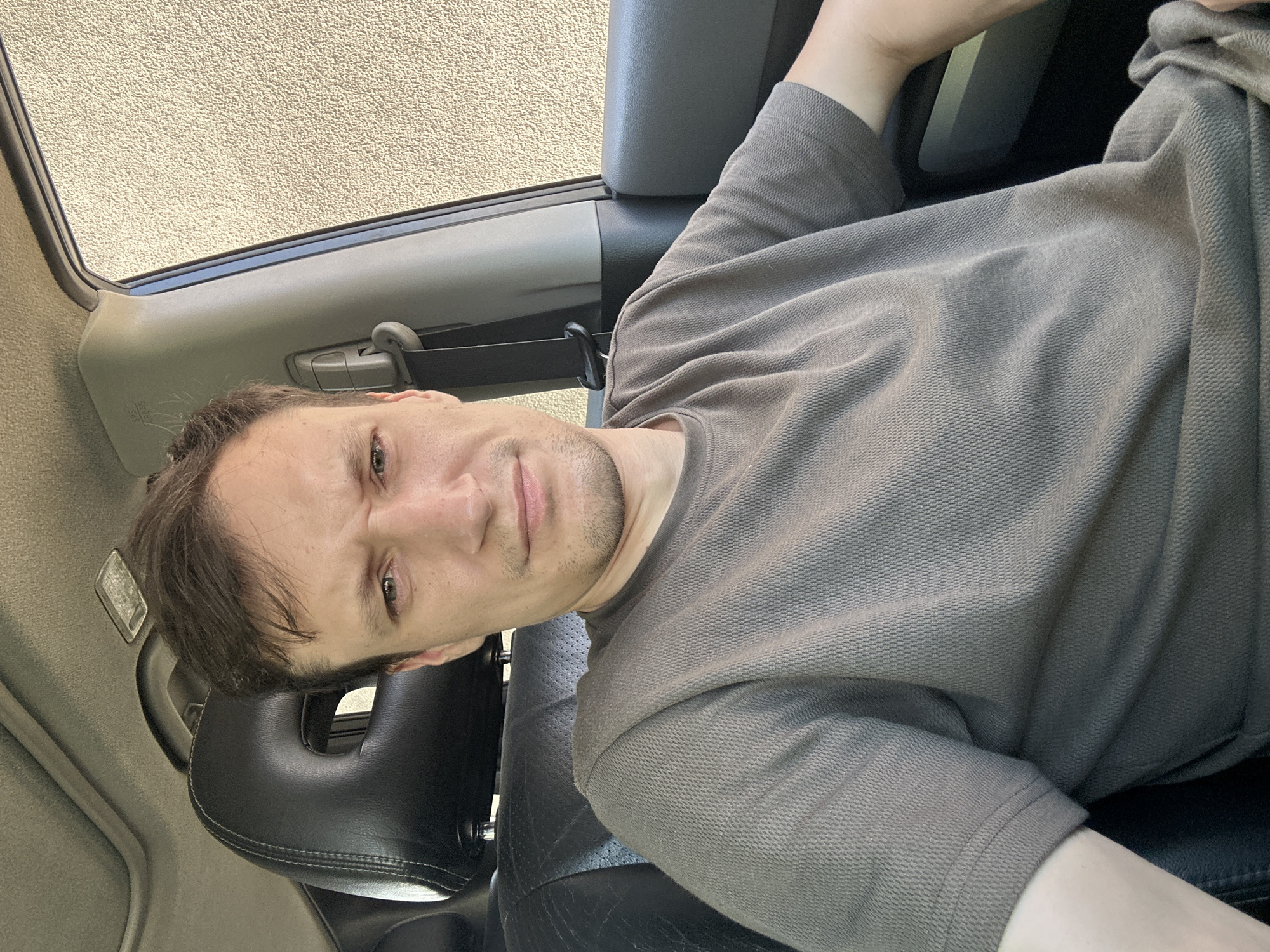 A medium shot shows a man sitting in the driver's seat of a car. He has light brown hair, a light complexion, and is looking directly at the camera with a serious expression. He is wearing a gray t-shirt. The car's interior is visible, including a black leather headrest with a circular cutout, the car's roof with a sunroof, and a gray dashboard. The man's right arm is resting on the car door. Outside the car, a textured, light-colored wall is visible through the window. The location is not specified within the image itself, and there is no information to suggest it is in Shatava, Ukraine.