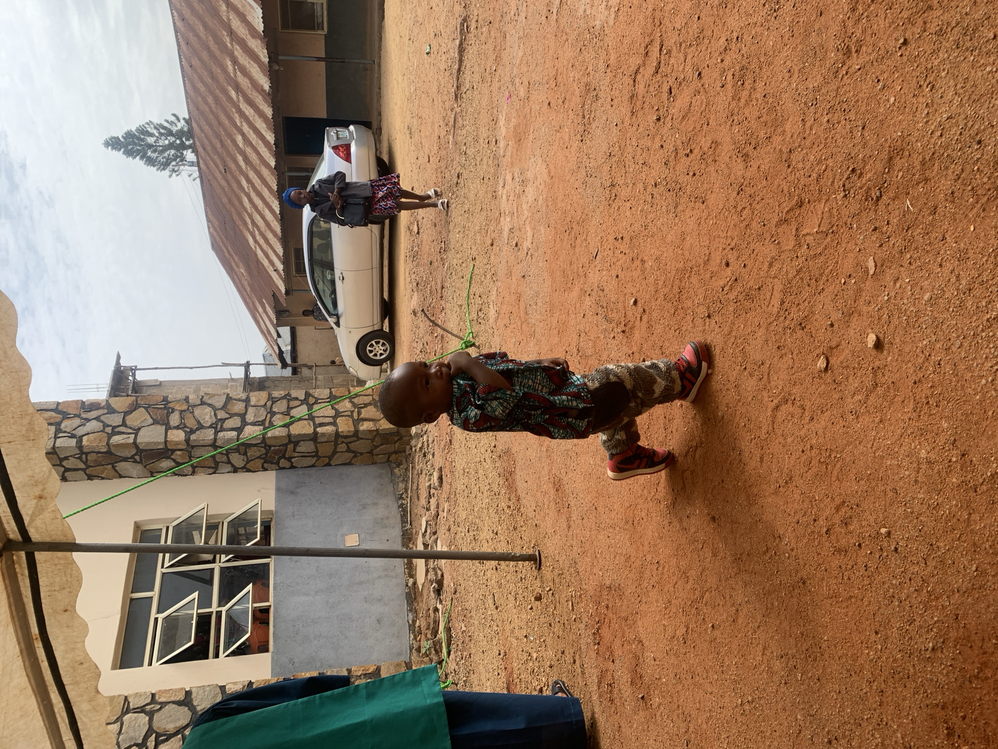 The scene shows a young child wearing patterned clothing and red sneakers, standing on a red dirt surface, possibly a yard. A thin green rope stretches across the frame horizontally. In the background, there is a white sedan parked next to a building with a corrugated metal roof. Further back, an adult stands near the car. In the foreground, the corner of a stone building with white-framed windows is visible. Part of a tent structure is also in view, indicating an outdoor event or gathering. Given the context of Jos, Nigeria, the red dirt surface is characteristic of the region.