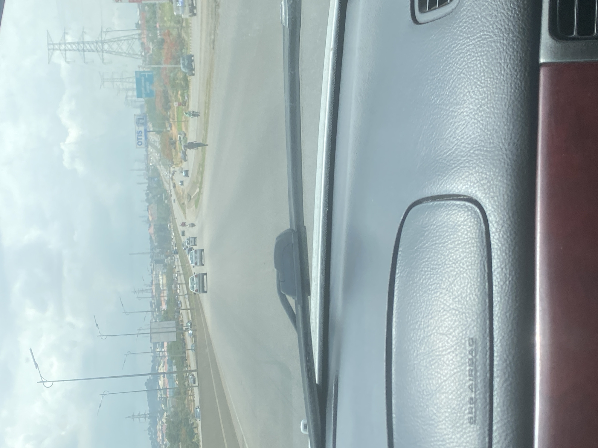 The image shows a view from inside a vehicle, likely a car, looking out onto a highway in Iddo Tudunwada, Nigeria. The dashboard, including an airbag panel, is visible in the foreground. Through the windshield, there is a clear view of a multi-lane highway with light traffic. Buildings and other structures are visible along the side of the road, including an "OTIS" sign and power lines. The weather appears overcast or hazy, obscuring some details of the background.