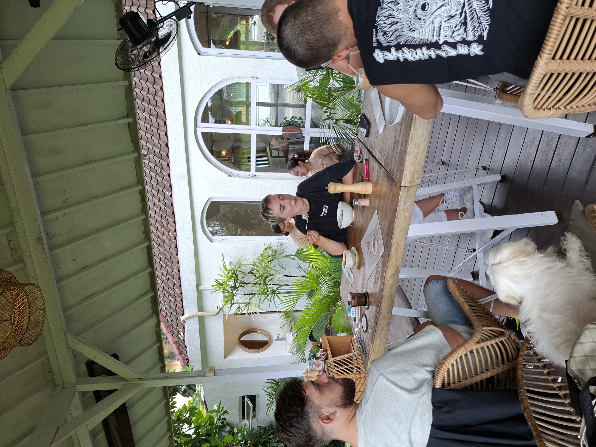 The image shows an outdoor dining area, likely in Ubud District, Indonesia. Several people are seated around a long wooden table, appearing to be eating and drinking. The woman wearing a black T-shirt with a logo that says “be yourself, because original is always better” appears to be sitting on a white chair and stirring a white cup on the table. The man sitting next to her has medium-length hair, a black shirt, and is drinking from a cup. A person is seated in the foreground wearing a gray shirt and blue shorts. A white dog stands near their chair. Tropical plants are visible around the dining area, and the architecture features white walls and arched windows. There is a ceiling fan hanging above the table.