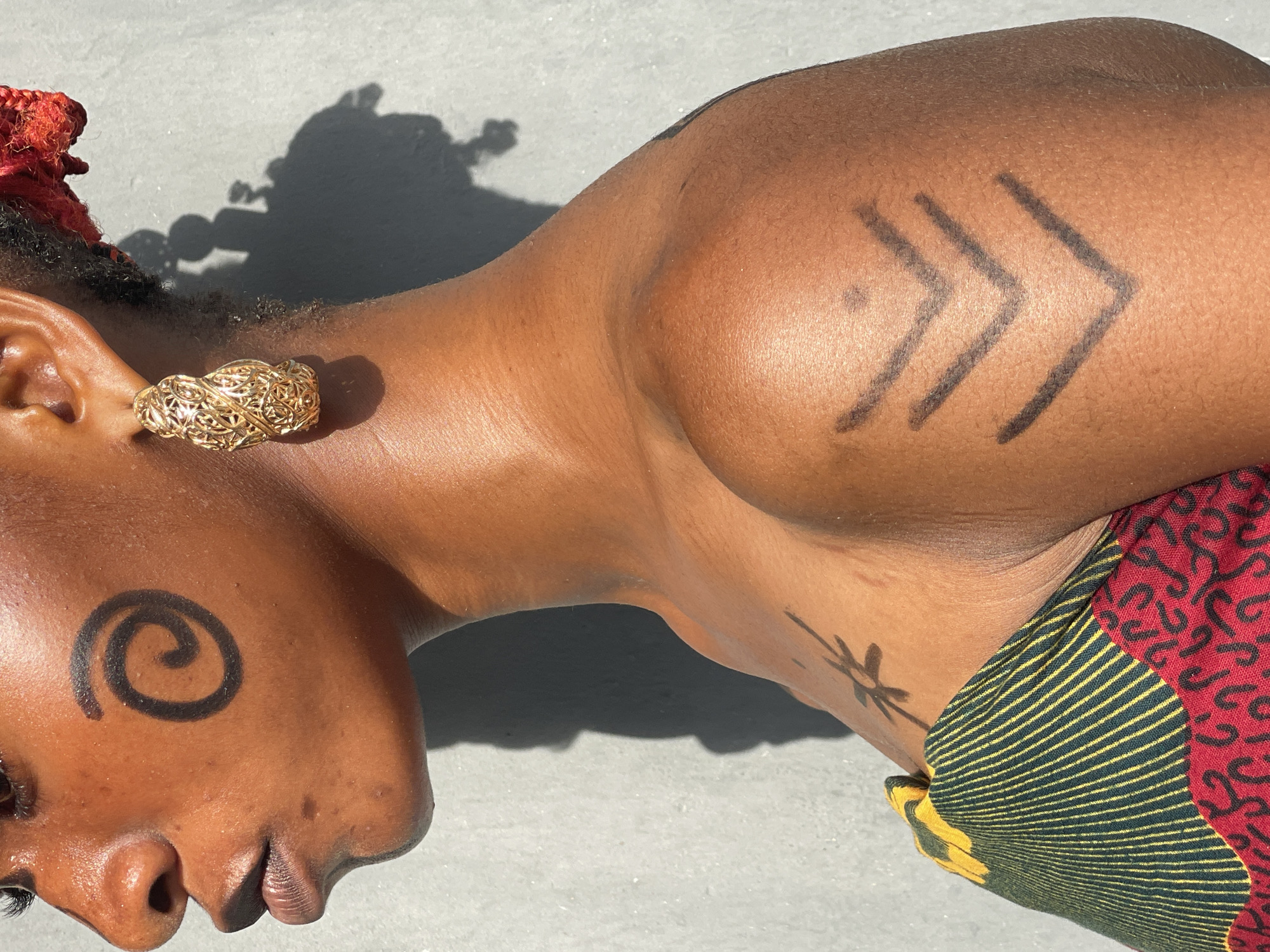 A person with dark skin lies on a light-colored, rough surface, possibly concrete. The person's head is turned to the left. On the left cheek is a drawn black spiral. The person wears a gold-colored, intricate earring on their left ear. The hair is styled with red fabric. Three black arrow symbols are drawn on the right upper arm, pointing to the right. A small black drawing of a sun or flower is on the neck. A red, green, and yellow patterned fabric is visible on the right side of the body. The image does not contain enough information to verify that it was taken in Badagry, Nigeria.