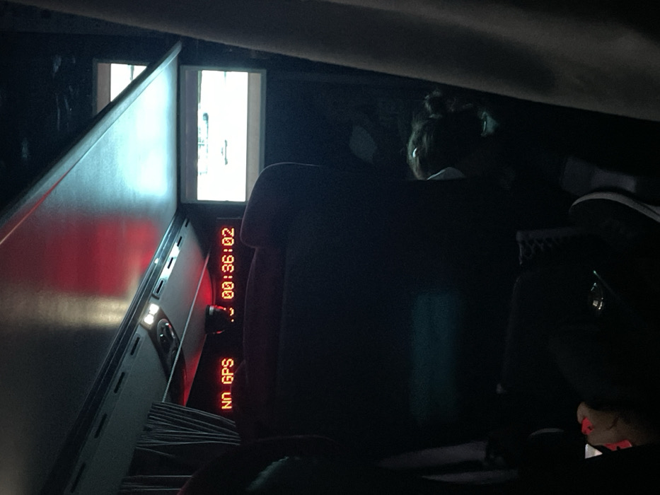 Inside a dark vehicle, a woman with a bun hairstyle sits facing forward. A "NO GPS" alert and a digital timer display "00:36:02" are visible on a console. A window or opening emits bright light. A portion of the vehicle's interior appears on the left side, illuminated by red light. The overall scene is dimly lit, obscuring many details.