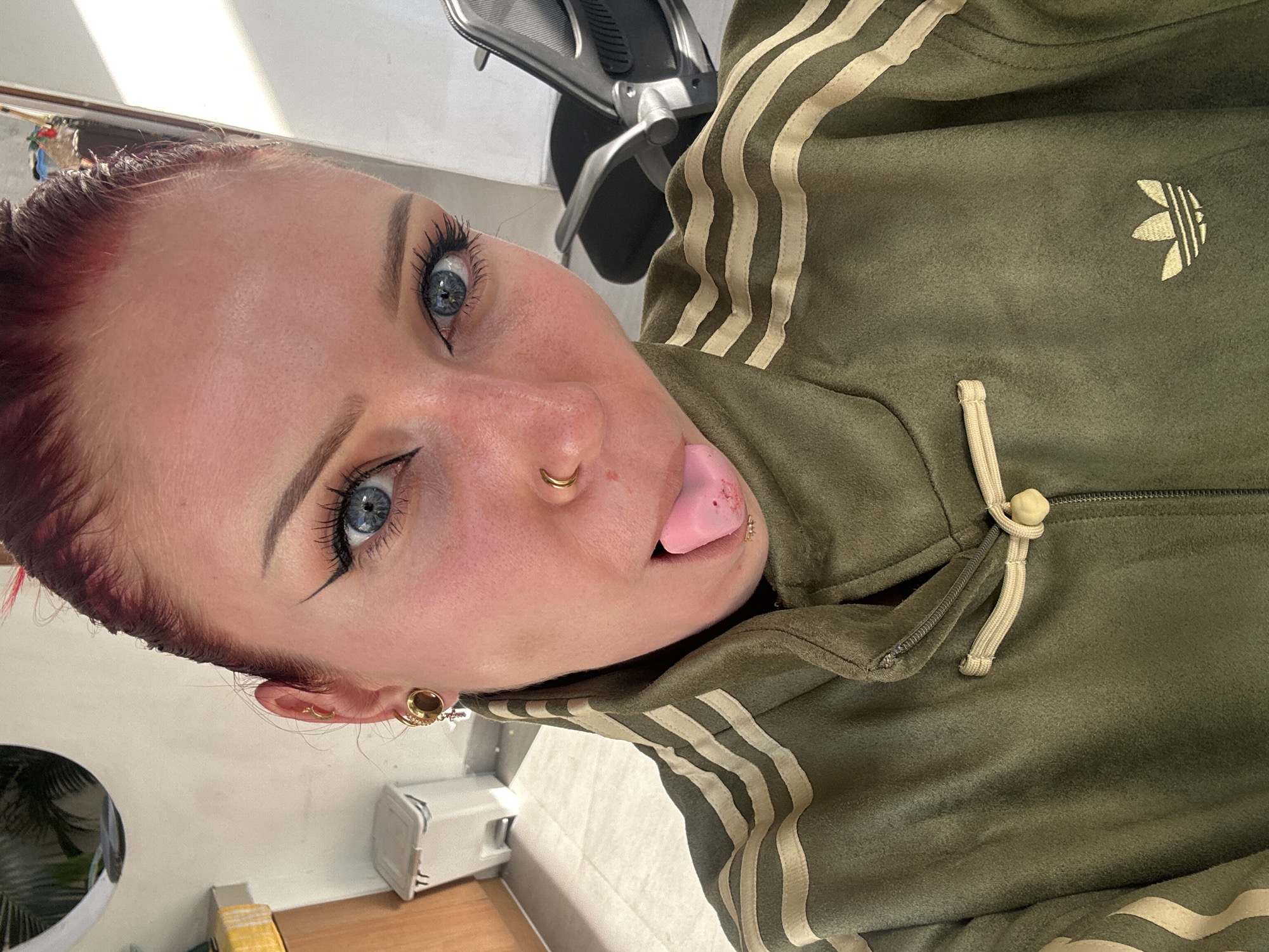 A close-up, tilted view shows a fair-skinned person with light blue eyes, dyed reddish-purple hair, and a nose piercing wearing a moss-green Adidas tracksuit top. The person's tongue is extended, and a gold stud earring is visible in the ear. In the background is a partial view of an office chair, a white object on a light surface, and a glimpse of a plant in a circular container. There is no evidence to suggest this image was taken in North Kuta, Indonesia.