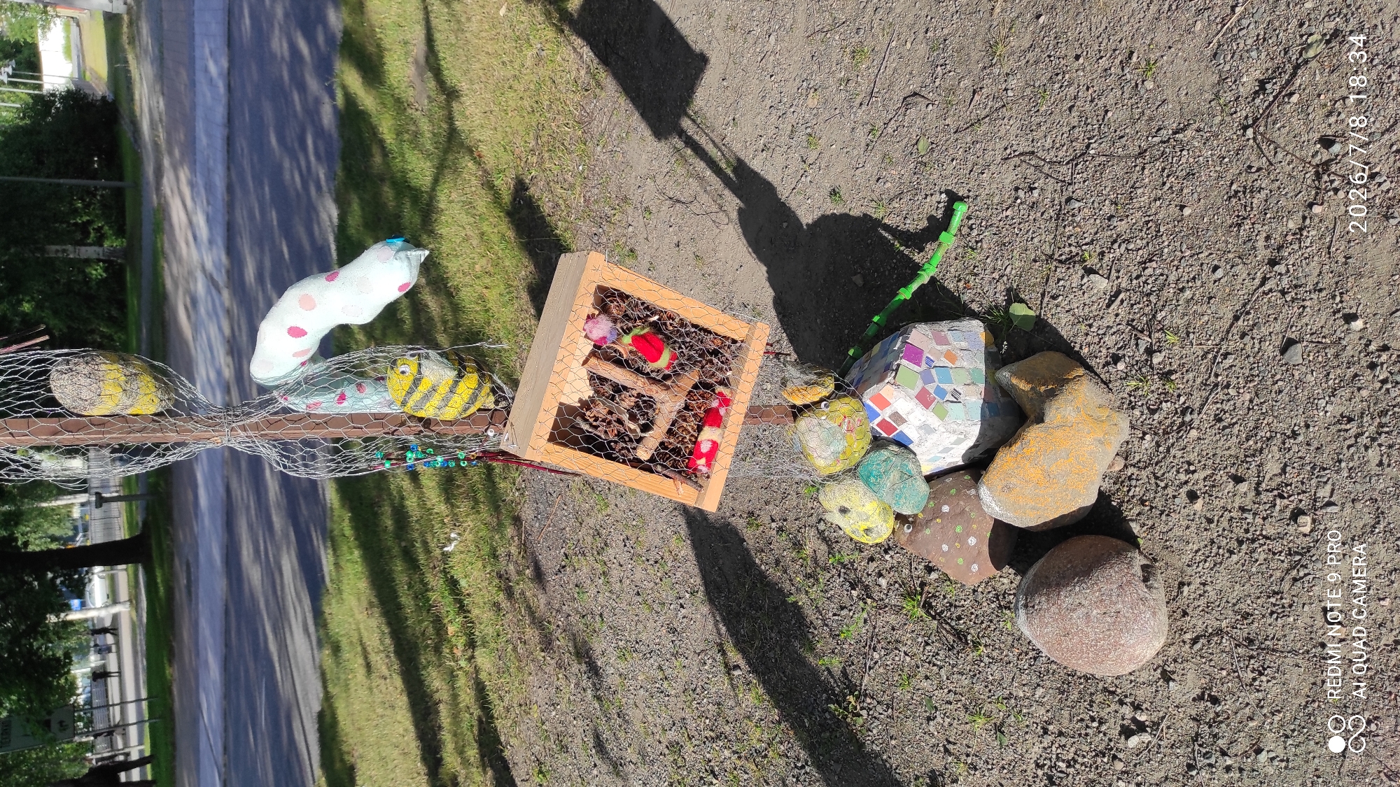 The image shows a decorative structure made of natural and craft materials, likely an art installation, situated outdoors in a public area with a paved road and grassy verge visible in the background. The structure features a wooden box filled with pinecones, possibly a shelter or feeder for small creatures. Surrounding the base of the structure are several decorated rocks, one of which is covered in colorful mosaic tiles. Suspended above the box by wire mesh are several fabric-like objects, including a yellow and black striped one resembling a bee, and a white object with pink polka dots. The scene is illuminated by sunlight, casting shadows on the ground. The timestamp in the lower right corner indicates the image was taken on July 8, 2026, at 18:34. The presence of streetlights, trees, and a road suggests an urban or suburban environment. Based on the style and context, this installation may be part of a public art initiative or a local community project in Iisalmi, Finland, potentially located in a park, along a walking path, or near a community center.