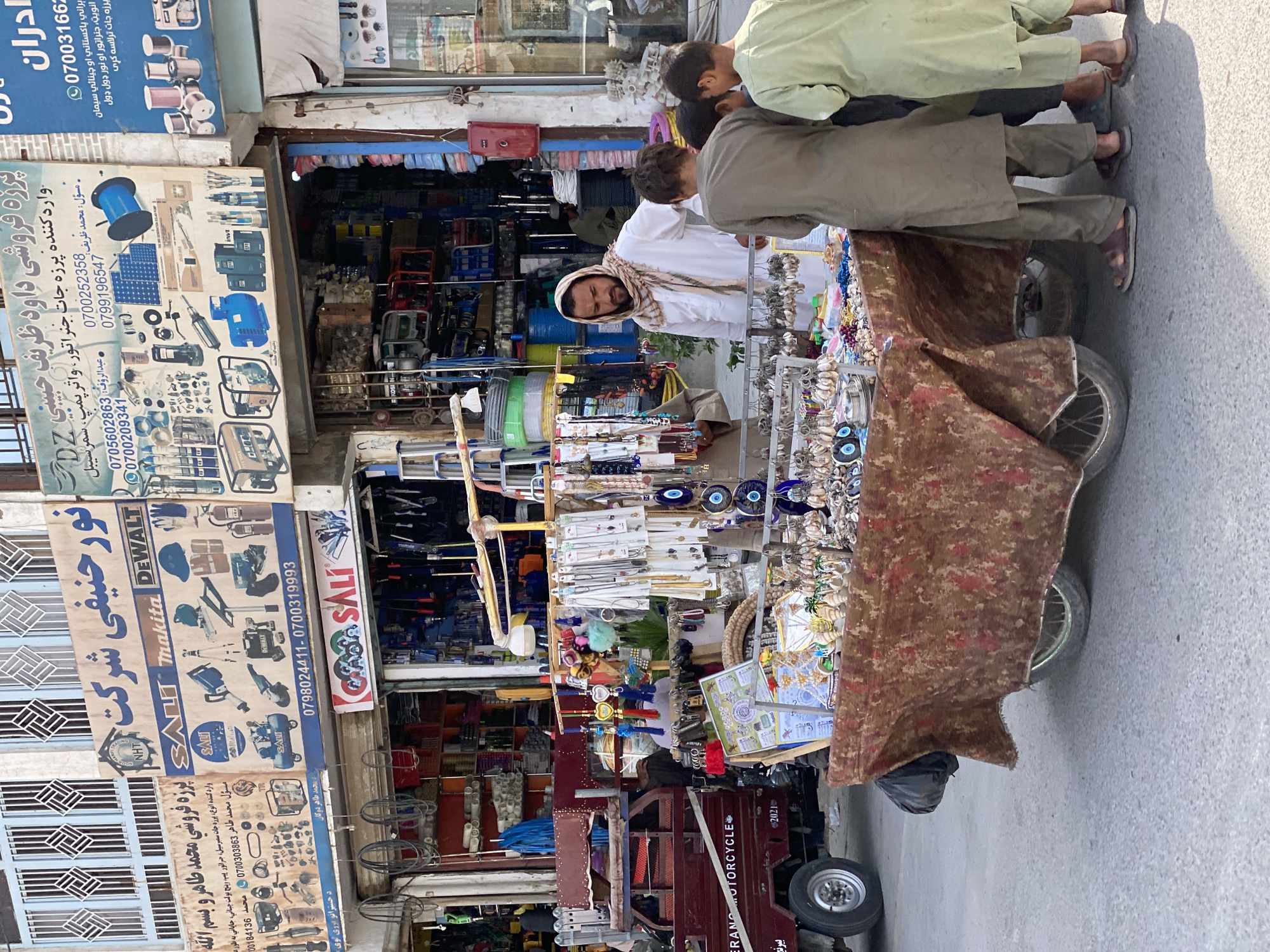 In the city of Kandahar, Afghanistan, a street scene unfolds in front of a shop selling hardware and electrical supplies. A man, wearing a white shalwar kameez and a patterned scarf, stands behind a mobile cart laden with various items. He appears to be interacting with a group of young boys who are gathered around the cart, looking at the merchandise. The cart is covered with a patterned cloth and displays an assortment of trinkets, necklaces, keychains, and other small decorative objects, including several evil eye charms. Behind the cart and the man, the shop is visible, with shelves packed with tools, generators, pumps, and other industrial equipment. A red truck with "MERCIAZO MOTORCYCLE" printed on its side is parked to the left of the scene. The overall impression is one of a bustling market environment with commercial activity and pedestrian traffic.