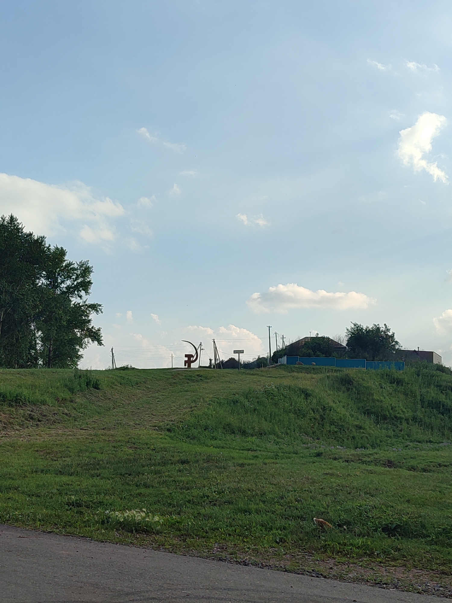 An outdoor, daytime shot captures a grassy hill under a partly cloudy sky. On the crest of the hill, a stylized sculpture resembling a sickle and hammer stands as a prominent landmark. In the distance, a few houses and utility poles are visible. A large green tree is situated on the left side of the frame. The foreground features a dark asphalt road. The scene is likely located in the village of Karabashyevo, Russia, given the presence of a sickle and hammer sculpture, which is a common symbol in rural Russia.