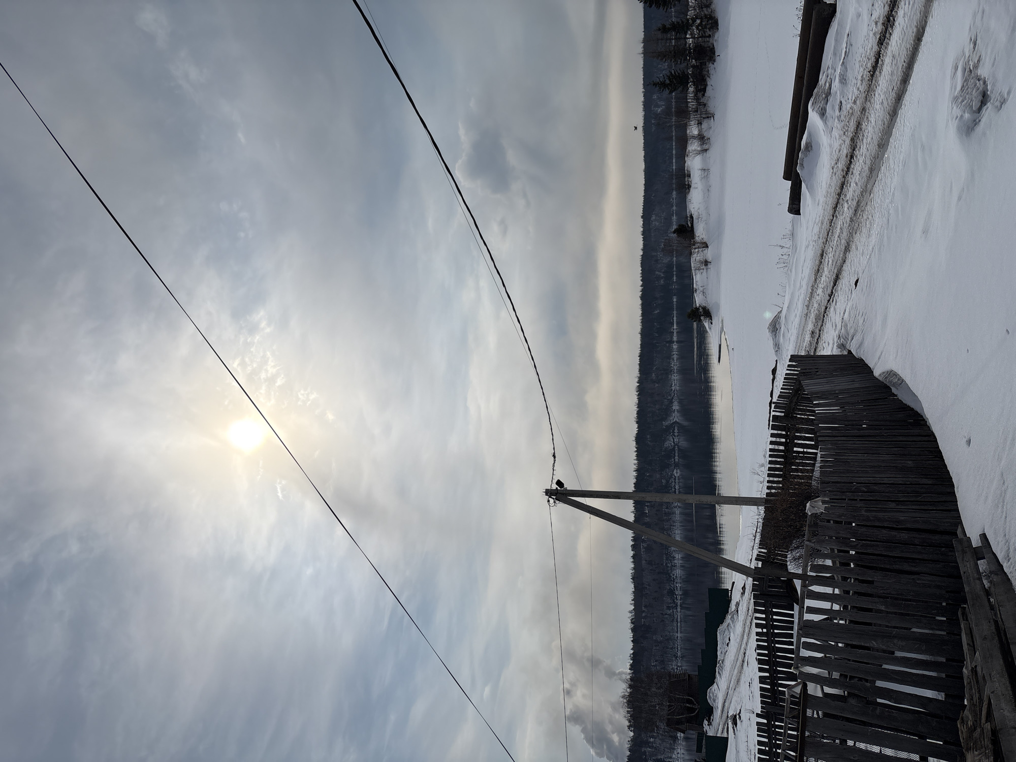 The image displays a tranquil winter landscape, likely set in Невон, Russia. The sky is partly cloudy, with the sun appearing as a bright, diffuse circle in the upper left quadrant, partially veiled by light clouds. Several dark power lines stretch across the frame, with a prominent one running diagonally from top-left to bottom-right. A wooden utility pole, situated towards the right, supports these lines, and a small, dark bird is perched on one of the wires near it.
The foreground on the right is dominated by a substantial layer of snow, forming drifts and covering a track or path. A rustic, weathered wooden fence runs along this snowy expanse. Beyond the fence and snow-covered ground lies a calm body of water, possibly a river or lake, which appears either frozen or extremely cold, reflecting the subtle hues of the overcast sky. The distant bank across the water is densely forested with dark evergreen trees. The scene conveys a sense of stillness and the stark beauty of a cold, quiet winter day. It is daytime, characterized by the visible, though diffused, sunlight. No human activity or visible text is present.