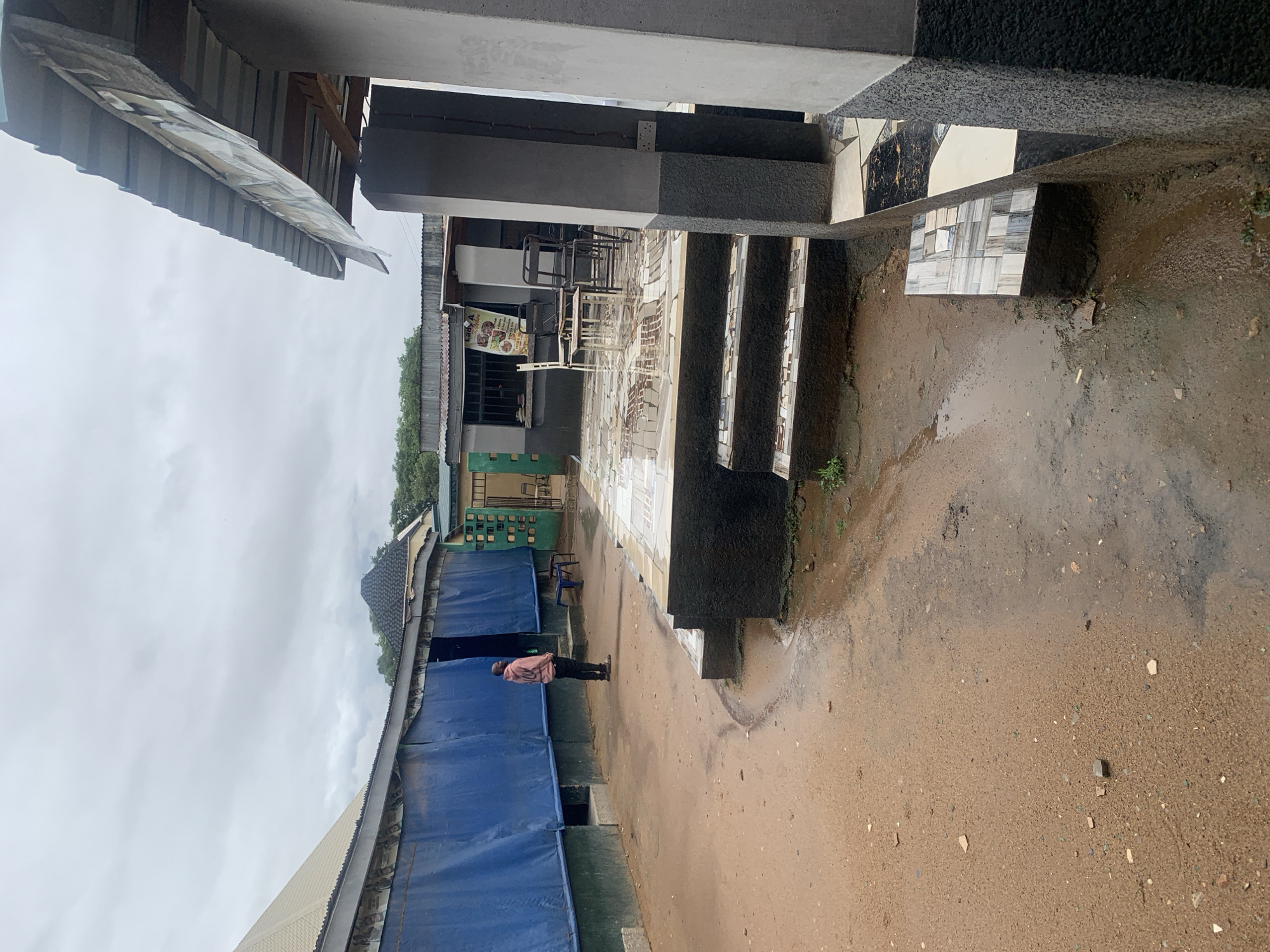 The image captures an outdoor or semi-outdoor commercial area in New Stadium, Nigeria, on an overcast day, evident by the cloudy sky and wet, muddy ground with puddles.
To the right, a concrete structure features pillars and steps, partly adorned with black, white, and grey tiles. Beyond these steps, a paved area with white and grey tiles serves as a dining space, furnished with several simple metal tables and chairs, some stacked. A vertical sign, likely a menu, is prominently displayed here. The visible text on the sign reads "LADY A..." at the top, followed by a list of food items including "FRIED RICE," "JOLLOF RICE," and "NOODLES," indicating a food establishment.
On the left side of the frame, several modest structures with corrugated metal roofs and blue tarpaulin walls line a path. A single individual, a man wearing a light-colored shirt and dark trousers, stands on this wet dirt path, facing towards the right side of the scene.
The general scene depicts a functional, informal setting, possibly a local eatery or food court, characterized by utilitarian construction and the aftermath of rain.