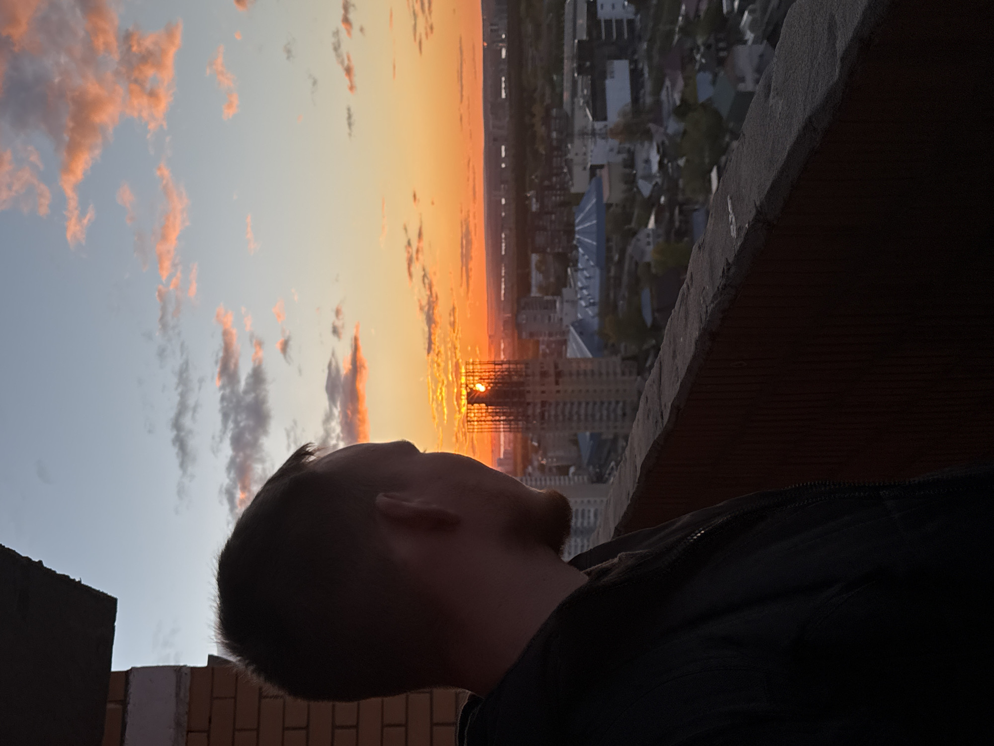The image depicts a man standing on a high balcony or rooftop overlooking a city at sunset. The man is in the lower center and right side of the frame, facing toward the sunset. The city, possibly Perm, Russia, stretches out in the background, with buildings and a mix of urban and green spaces visible. The sunset is characterized by a gradient of orange hues bleeding into the sky, partially obscured by buildings. The edge of the balcony or rooftop, constructed from brick and concrete, is in the foreground.