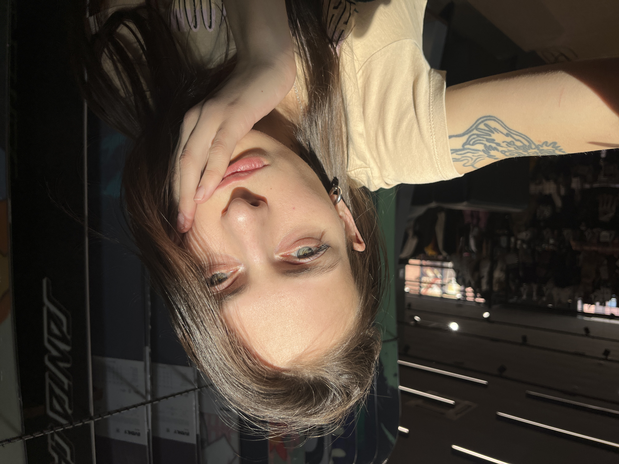 A woman with long brown hair, green eyes, and a light complexion poses for a photo. Her right hand partially covers her face. She wears a beige t-shirt and a hoop earring on her left ear. A tattoo is visible on her right arm. The background appears to be a store interior with shelves and merchandise. Light fixtures are visible on the ceiling. The scene is located in the city of Krasnoyarsk, Russia.