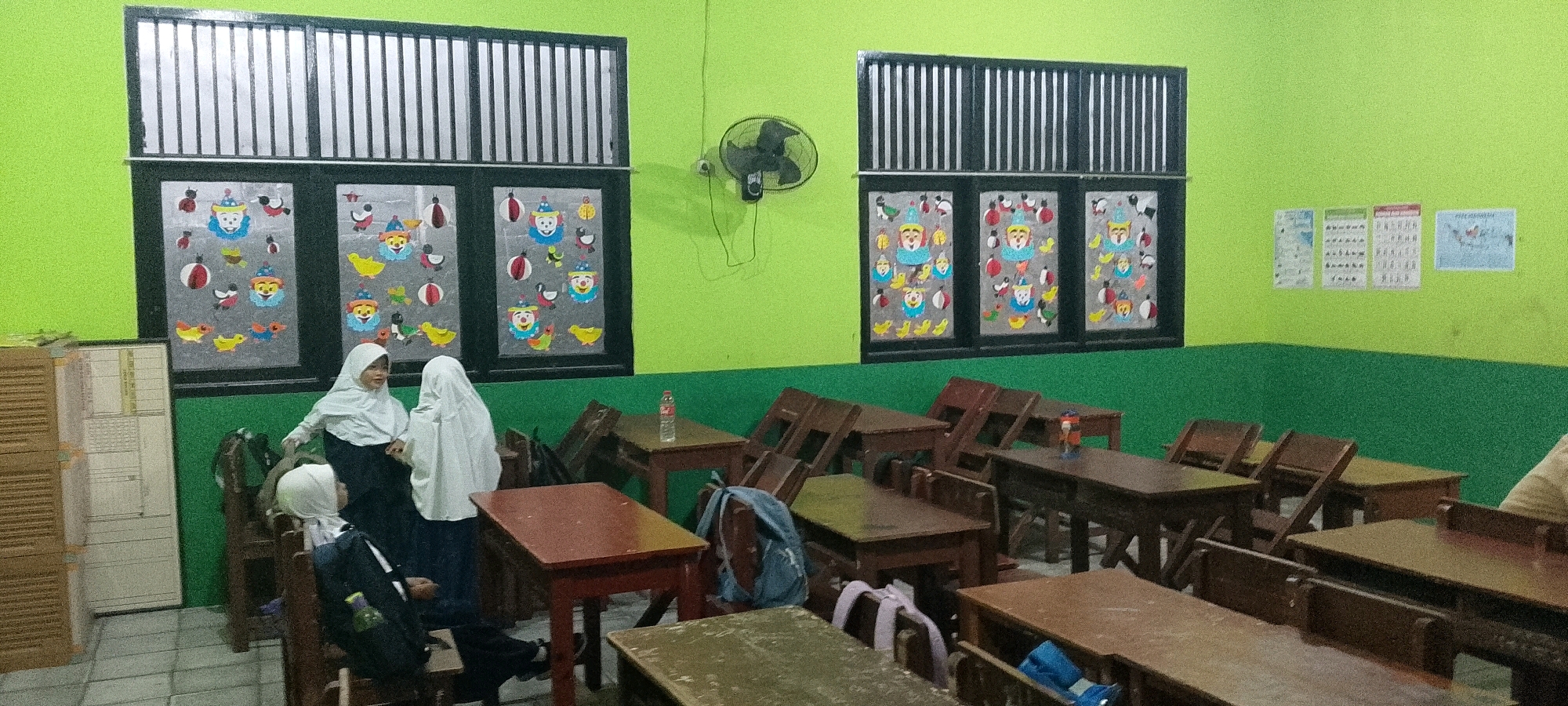 The image depicts an indoor scene, likely a classroom, in Satriajaya, Indonesia. The room has light green walls and a darker green baseboard. Two large windows with black frames and vertical blinds are visible at the top of the walls. Below these windows are smaller window panes decorated with colorful cartoon cutouts, primarily of clowns and animals like ducks and birds.
In the foreground and midground, several wooden desks and chairs are arranged in rows, some pushed together. The desks appear to be made of dark wood, and some have a reddish-brown finish. A water bottle is placed on one of the desks. A backpack and other items are visible on and around the desks.
On the left side of the room, there is a tall, light-colored cabinet with horizontally slatted doors. On the wall to the right of the windows, there are several posters or charts. A black oscillating fan is mounted on the wall.
Three individuals, all wearing headscarves, are present in the scene. Two are standing near the windows on the left, and one is seated at a desk in the midground. They are dressed in dark clothing, suggesting they might be students.
The overall impression is of a functional, possibly elementary school, classroom environment in Satriajaya, Indonesia, decorated with child-friendly imagery.