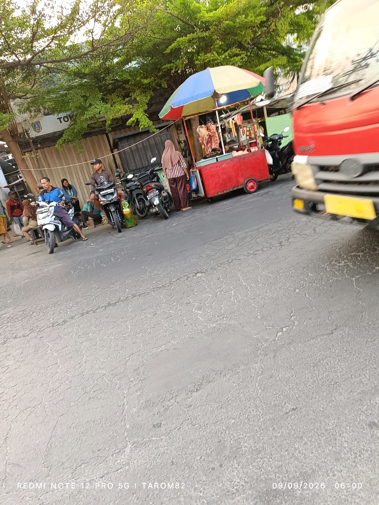 The image captures a street scene in Ngoro, Indonesia, on a sunny day. In the foreground, a red truck is partially visible, its front bumper and headlights in focus. The road surface is asphalt, showing signs of wear and tear with a cracked texture. To the left, a street vendor's stall is set up under a colorful umbrella, displaying various cuts of meat. Several motorcycles are parked near the stall, with a few people gathered around. One man in a blue shirt is sitting on a white motorcycle, while another man in a brown shirt and cap is on a black motorcycle. A woman in a hijab and striped shirt is standing near the stall. The background features buildings and lush green trees, indicating an urban environment. The bottom of the image contains text displaying "REDMI NOTE 12 PRO 5G | TAROM82" and a date and time stamp of "09/09/2026 06:00".