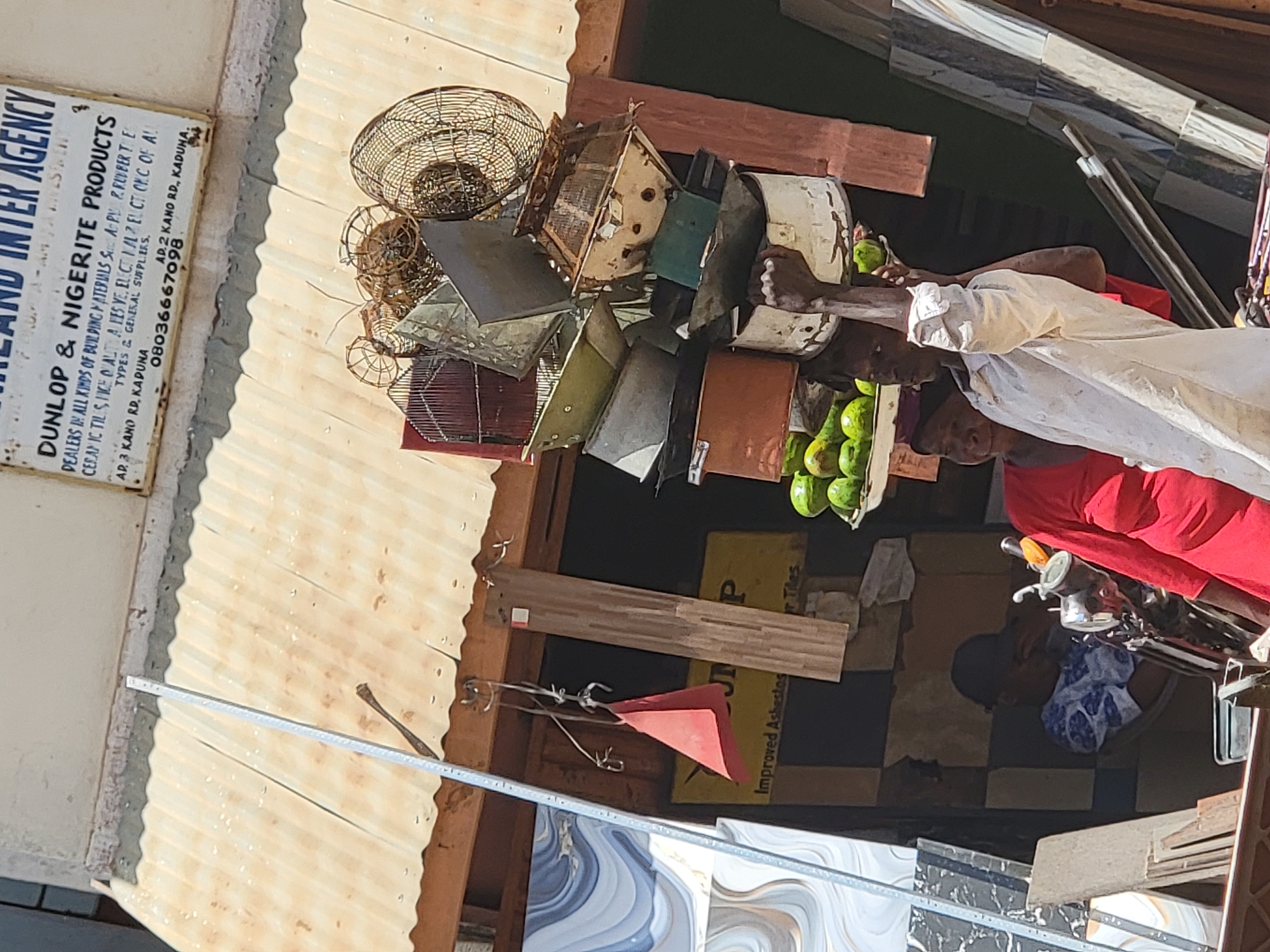 The image shows an outdoor market scene. A middle-aged woman in a red shirt and white wrap is selling what appears to be green fruit (possibly avocados). The market is located under a corrugated metal roof. A sign reading "DUNLOP & NIGERITE PRODUCTS" is visible, indicating the sale of building materials. Other objects include metal cages, pots, and tiles. A man in a blue and white shirt can be seen in the lower part of the image. A motorcycle is parked nearby. The sign indicates the location to be along Kano Road, Kaduna. No information is found about the location being in the city of Maiburiji.