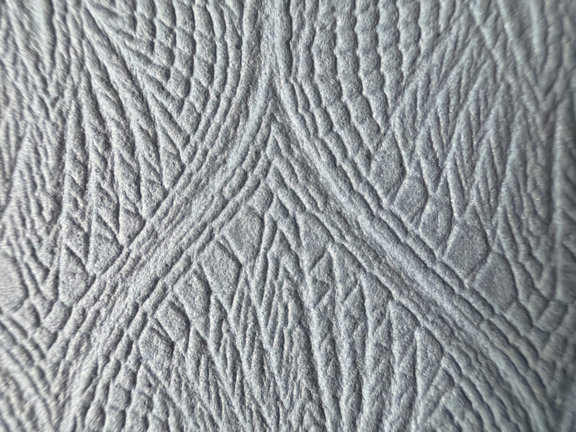 The image shows a close-up of a textured, gray surface. The texture consists of a repeating pattern of raised lines and depressions, creating a visual effect similar to wrinkled skin or crumpled paper. The lines are arranged in a somewhat organized manner, with converging and diverging patterns that form triangular shapes. The lighting appears to be diffused, highlighting the texture without casting strong shadows. There is no evidence to link the image to Saint Petersburg, Russia, or to any specific location. Furthermore, the image contains no people, subjects, objects, actions, or interactions beyond the texture itself.