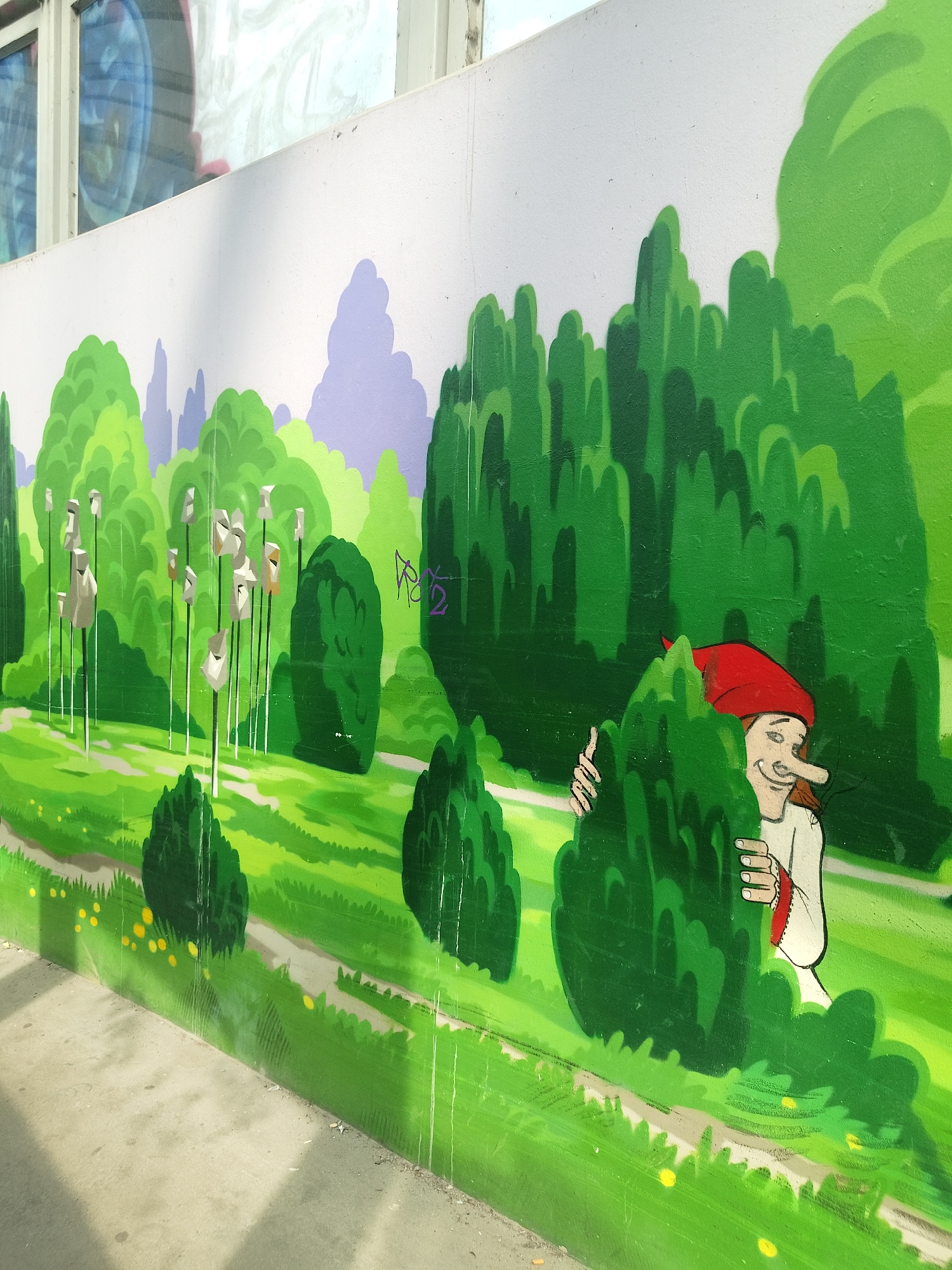 A vibrant mural adorns a concrete wall in an urban setting. The artwork depicts a whimsical landscape dominated by lush green foliage and stylized trees. A path winds through the scene, dotted with yellow wildflowers. Amidst the greenery, a cartoonish figure with a red cap peeks out from behind a bush, adding a playful element to the composition. The mural's location is Unknown, Unknown.