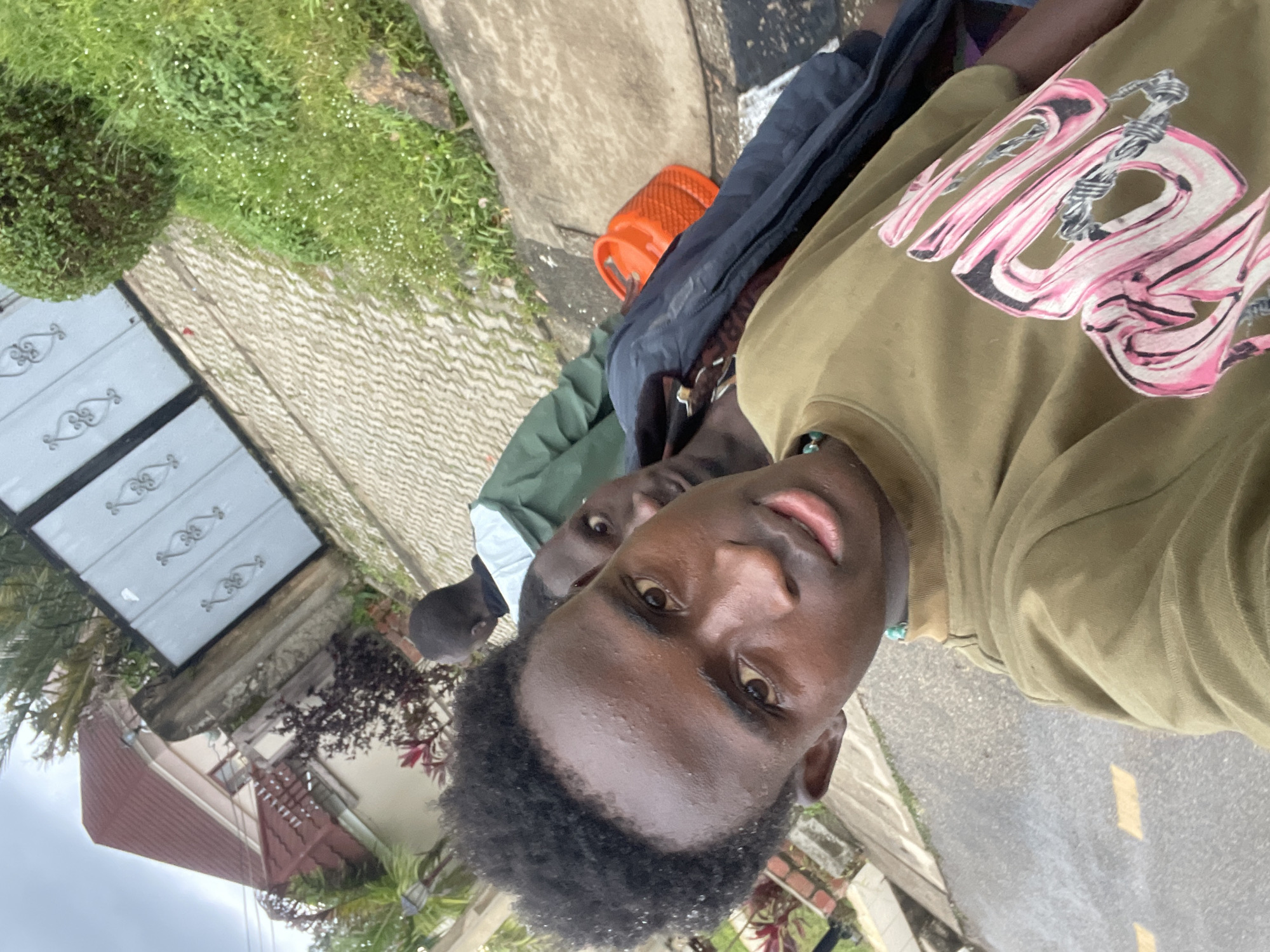 The image captures a selfie taken at an outdoor location, possibly in the city of Jos, Nigeria, given the context provided. The foreground is dominated by the face of a young Black male, looking directly at the camera. He has dark, curly hair and is wearing an olive-green t-shirt with a distinctive pink and black graphic design featuring barbed wire elements. A turquoise beaded necklace is visible around his neck.
Behind him, partially obscured, are at least two other individuals. One person is wearing a dark jacket or hoodie, and another is wearing a green hooded jacket. The background reveals a paved walkway or driveway with a herringbone pattern, leading to a metal gate with ornate scrollwork. Adjacent to the paved area is a patch of lush green grass with small white flowers and a large, rounded green bush. A stone is visible in the grass. Further in the background, a building with a red-tiled roof and light-colored walls can be seen, along with some reddish-purple foliage. An orange object, possibly a gas cylinder, is placed on the ground next to the paved area. The overall lighting suggests an overcast or diffused daylight.