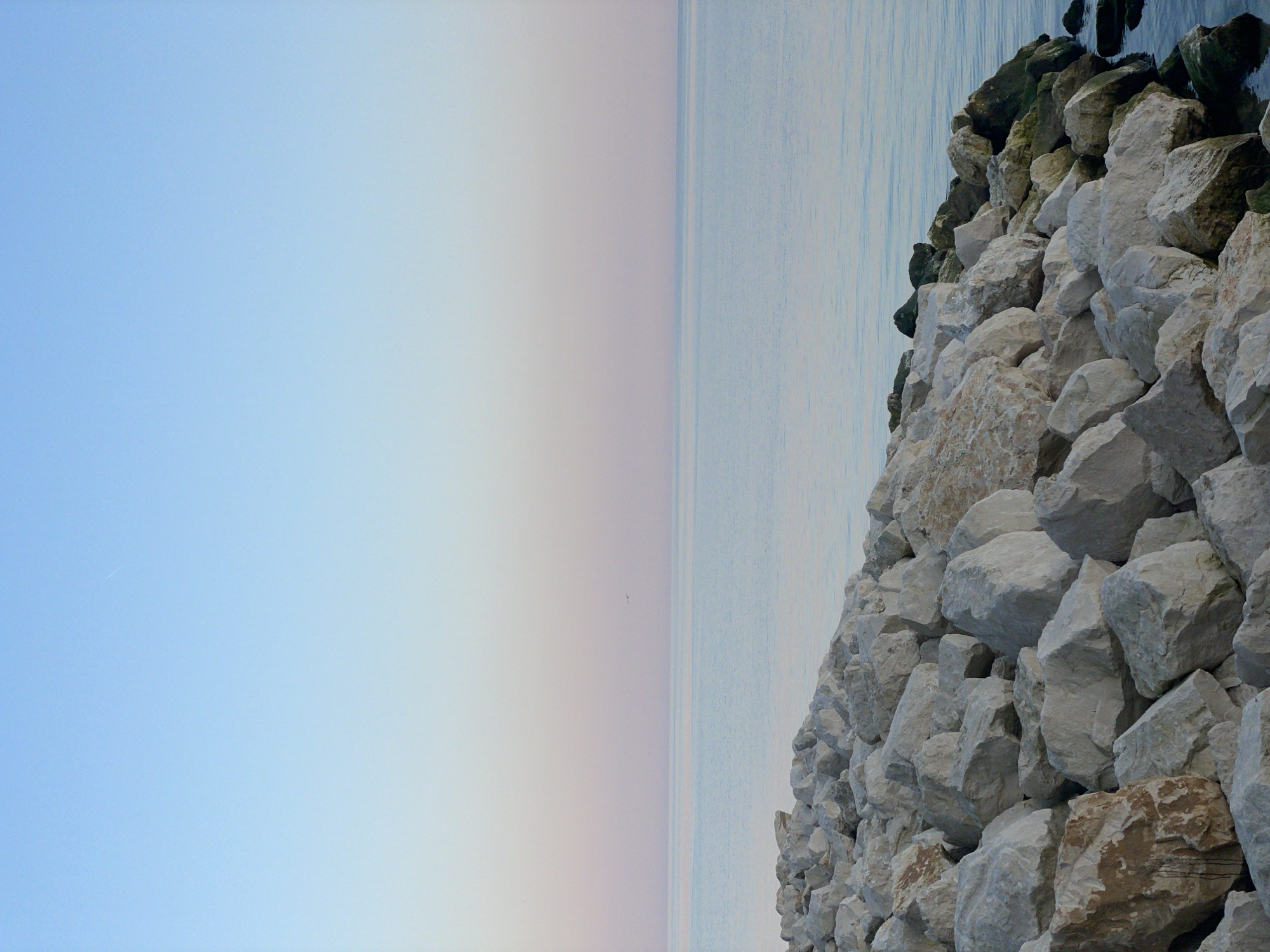 The image depicts a scene from Varna, Bulgaria, featuring a breakwater composed of large, roughly hewn white and gray stones on the right side. The stones are densely packed and appear to be protecting the shoreline. To the left of the breakwater, the image shows a calm body of water, likely the Black Sea, meeting a pastel-colored sky. The sky transitions from a light blue near the top-left to a soft pinkish-gray near the horizon, indicating either sunrise or sunset. The horizon line is straight and separates the sky from the sea. There are no visible people in the image. A small line of dark grey/black coloured rocks is on top of the breakwater. The stones in the breakwater appear to be in varying sizes, from small to quite large.