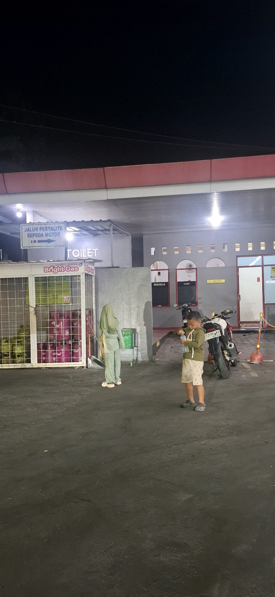 This image depicts an outdoor scene at night, likely at a gas station in Colomadu, Indonesia. In the foreground, a young boy, appearing to be around 5-7 years old, stands facing left, holding a clear plastic water bottle in his right hand and his left hand near his mouth. He is wearing a greenish-brown long-sleeved shirt and patterned shorts, with dark gray clogs on his feet. To his left, a person wearing a light green headscarf and matching long-sleeved outfit and pants stands facing away from the camera. This individual is wearing light-colored slip-on shoes.
Behind the boy and the person in green, several motorcycles are parked. One is prominently displayed in the middle ground, a white and red motorcycle with the license plate C 5388 YU. To the left of the motorcycles, a metal cage structure contains rows of pink and green gas cylinders. A sign above the gas cylinders reads "Bright Gas". A white sign with blue lettering indicates "JALUR PERTALITE SEPEDA MOTOR" with an arrow pointing left, and another sign in larger font reads "TOILET".
The environment appears to be paved, with a slight slope leading towards the buildings in the background. The buildings are illuminated by overhead lights. Structures visible include entrances, windows, and what appears to be a Musholla (prayer room) indicated by a sign. The overall impression is that of a functional, dimly lit public space at night.