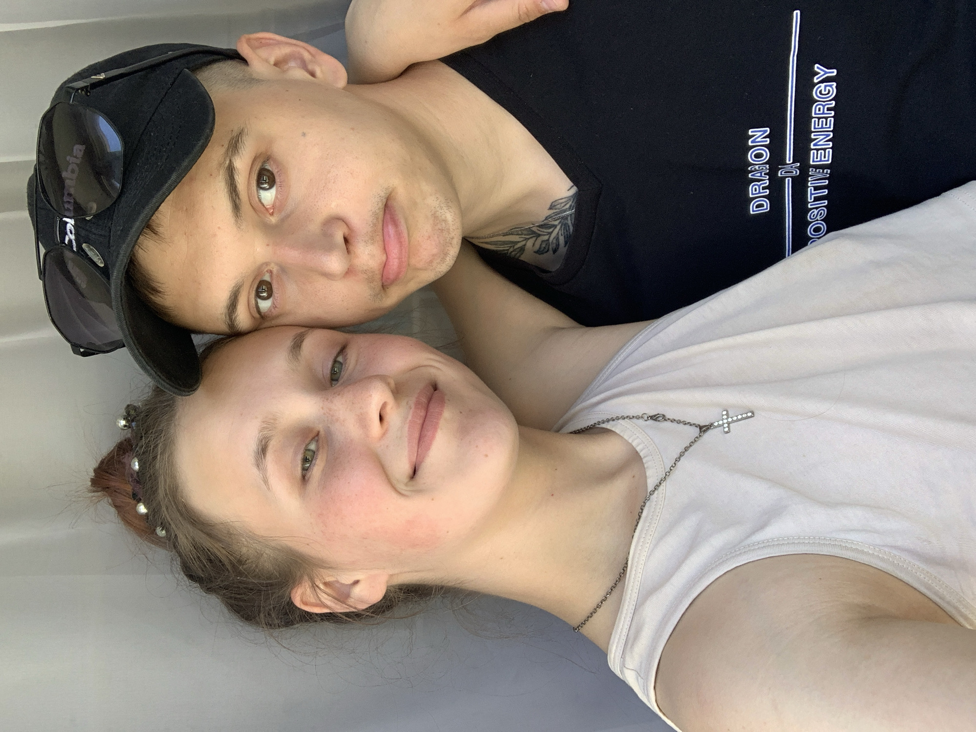 Two people are lying down, facing upward toward the camera. The person on the left wears a black baseball cap with sunglasses perched on top, and a black t-shirt with white writing on the right breast. The person on the right wears a light-colored t-shirt and a necklace with a cross pendant. They appear to be indoors, resting on a light gray surface. There are no elements to suggest that the photo was taken in the city of Фоки, Russia.