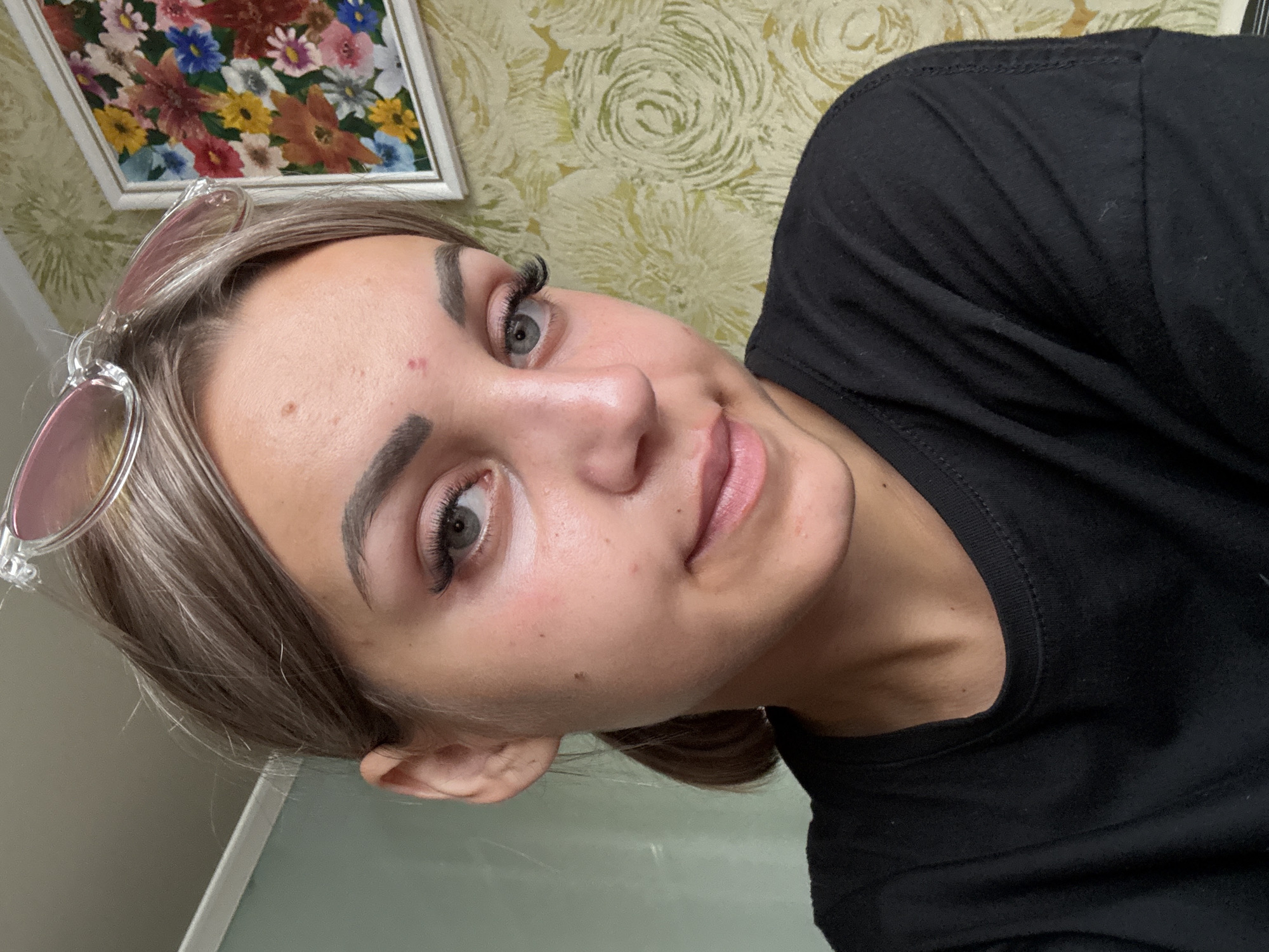 A young woman with blonde hair pulled back in a ponytail and wearing a black t-shirt is shown in a close-up portrait. She is looking at the camera with light blue eyes and a slight smile. She is wearing large false eyelashes and has noticeable makeup, including blush and lip gloss. A pimple is visible on her forehead, and another on her cheek. A pair of pink-tinted sunglasses are perched on top of her head. The background consists of a wall with a floral wallpaper pattern and a framed picture of flowers. The lighting suggests an indoor setting. The image was taken in Krasnogorskoye, Russia.