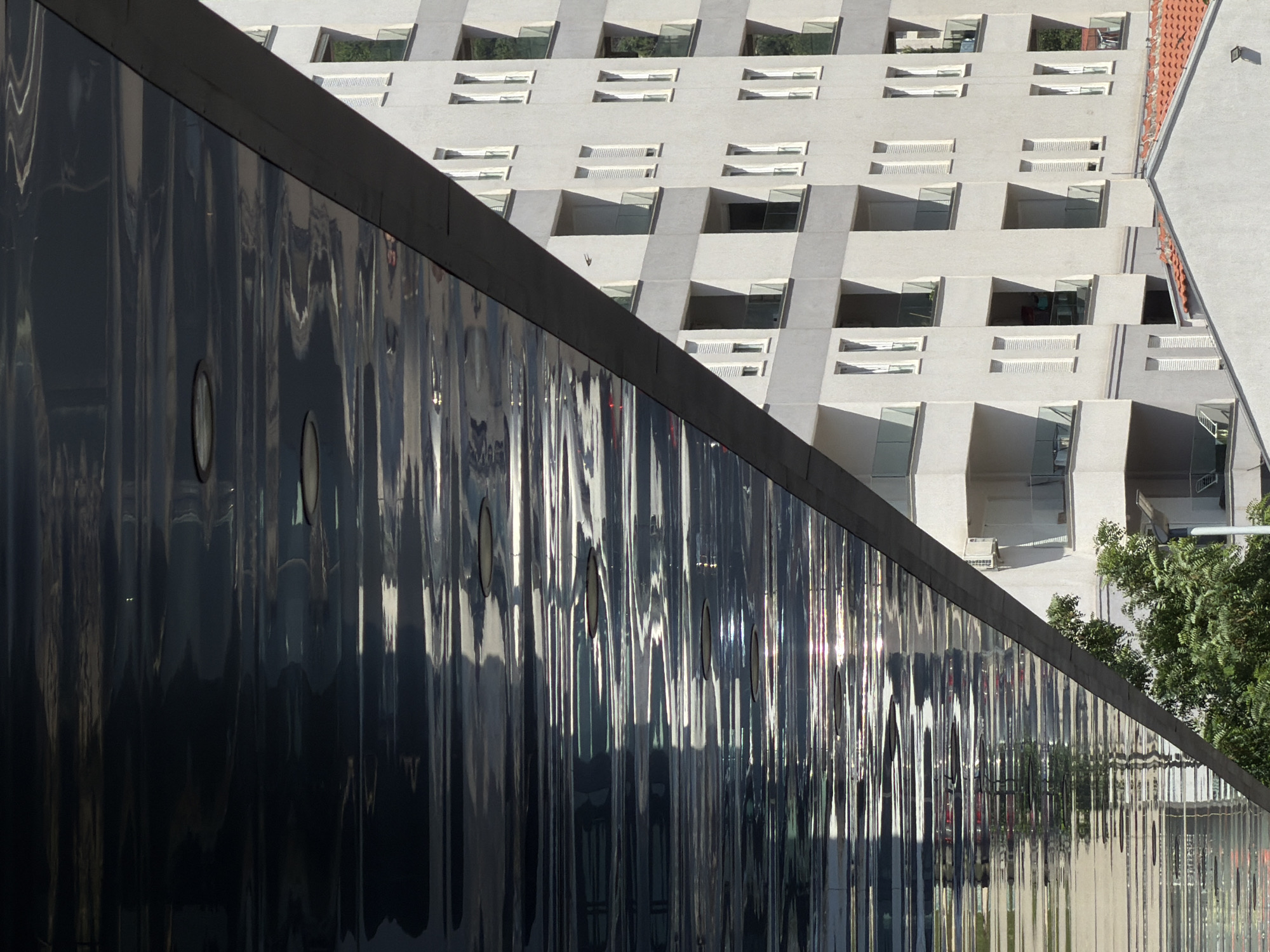 The image depicts an architectural scene likely in Budva, Montenegro, given the style and location. In the foreground is a reflective dark surface with small circular openings, possibly a modern architectural element. Behind it rises a light-colored building with a unique, stepped façade featuring rectangular windows and angled surfaces. A section of a terracotta tiled roof is visible on the upper right. Some greenery, likely trees, is present on the right side of the frame. The environment appears to be urban, with modern buildings and architectural designs prominent. There are no people visible in the scene.