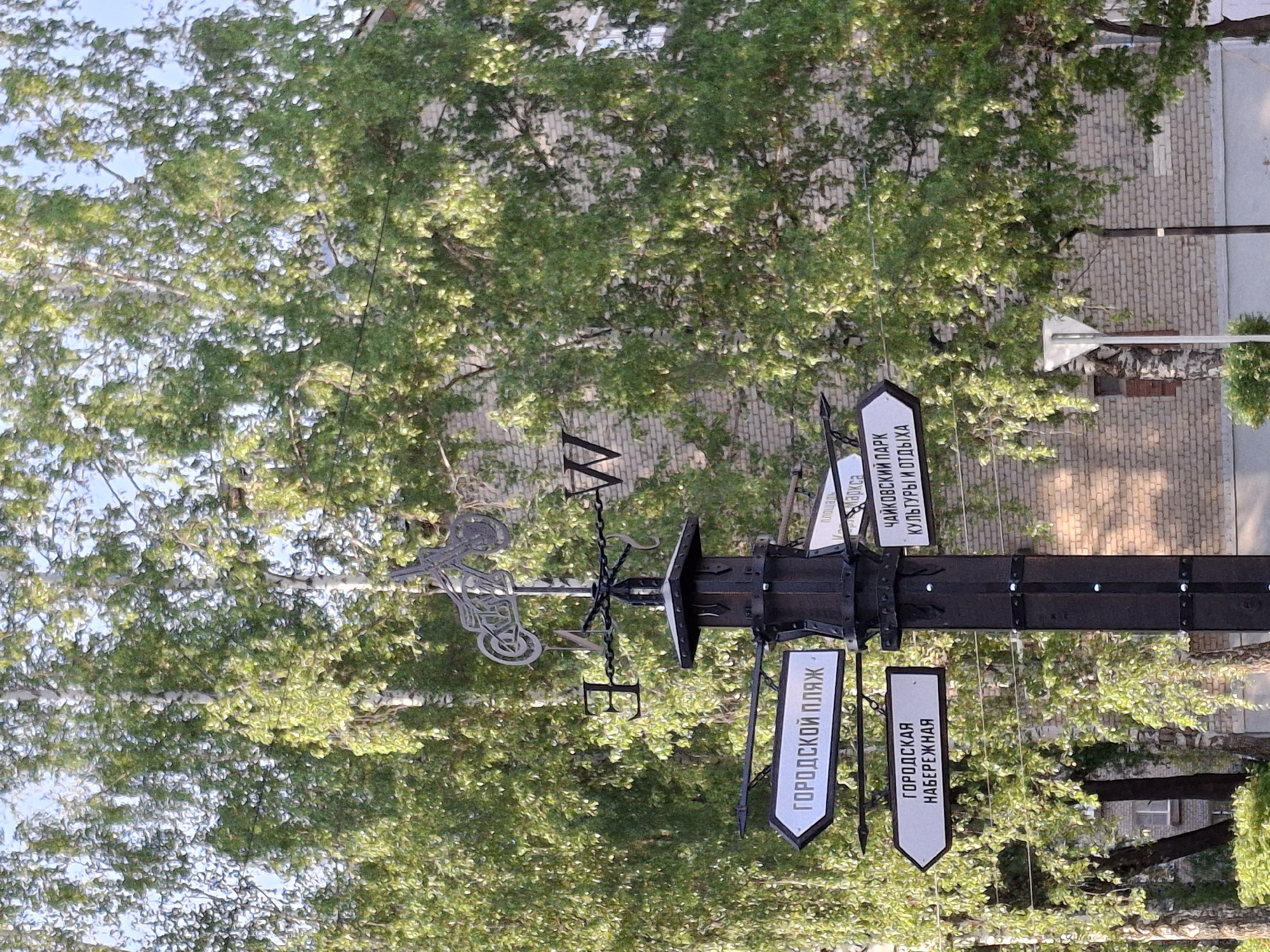 A black, metallic directional signpost, with an ornate weather vane on top, stands among trees in the city of Chaikovsky, Russia. The signpost features white signs with black text in Russian. One sign reads "ГОРОДСКОЙ ПЛЯЖ" (City Beach). Another sign reads "ГОРОДСКАЯ НАБЕРЕЖНАЯ" (City Embankment). A third sign reads "ЧАЙКОВСКИЙ ПАРК КУЛЬТУРЫ И ОТДЫХА" (Tchaikovsky Park of Culture and Leisure). The weather vane includes the cardinal directions "N," "E," "W," and "S" with an ornate bicycle and rider design. A brick building is visible in the background.
