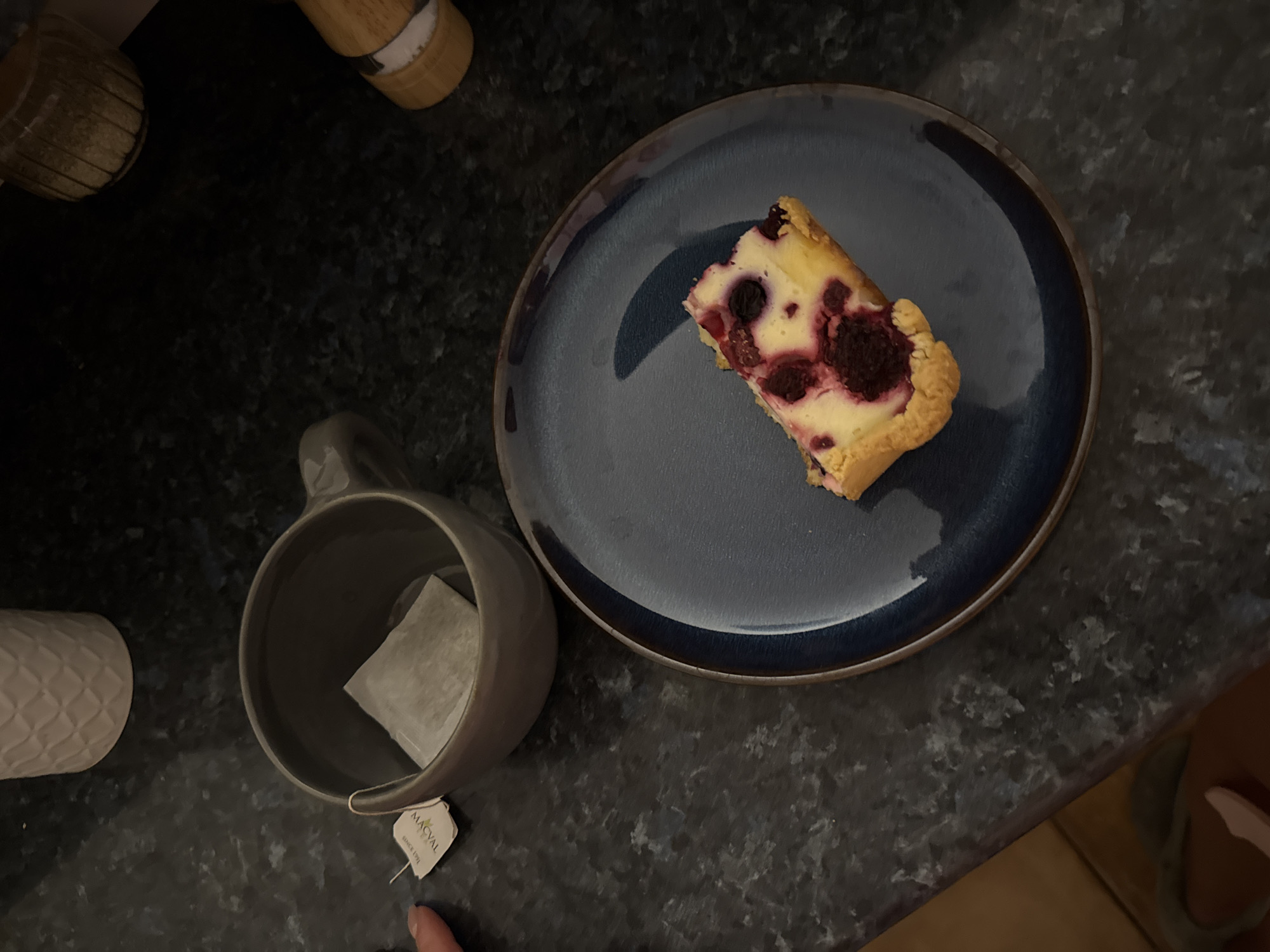The image shows a top-down view of a dark countertop with several objects. A slice of raspberry cheesecake sits on a blue plate. A gray mug containing a tea bag with the tag visible is to the left of the plate. Further to the left, there is a white patterned container and a wooden object. The countertop surface is dark and speckled. The scene appears to be set indoors. There is no evidence to determine this is the city of Zankovići, Montenegro.