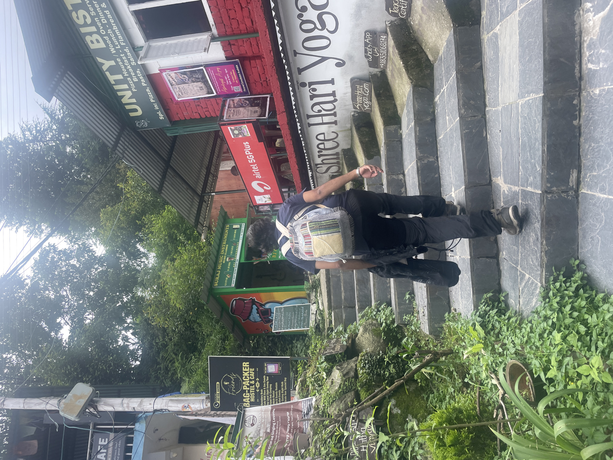 The image depicts a steep stone staircase in Bhagsu, India, leading upwards towards a cluster of shops and businesses. A person, seen from behind, is walking up the stairs carrying a backpack. The surroundings are lush with green foliage on the left side of the stairs, with several signs indicating various establishments. To the right, businesses like "UNITY BISTRO" and "Shree Hari Yoga" are visible, with additional signage related to their services and contact information. The overall scene captures a common street view in a hilly or mountainous tourist area.