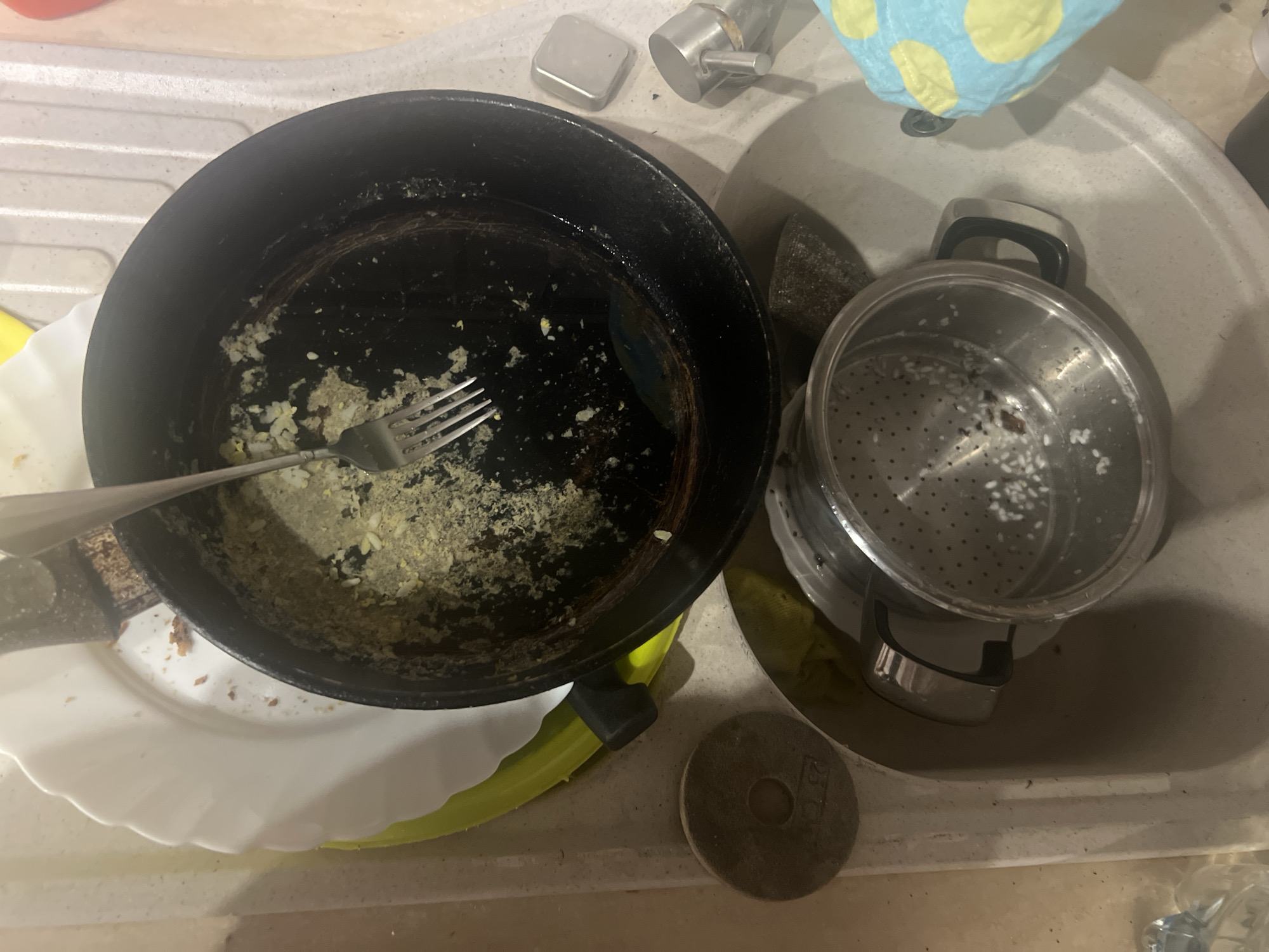 The image shows a messy kitchen sink in a dimly lit setting, possibly at night or indoors. The sink contains dirty dishes, including a frying pan with leftover food residue and a fork, a steamer pot with rice, and plates. There are also some cleaning supplies and a dish towel with a yellow and blue polka dot pattern. The overall scene suggests that a meal has recently been prepared and the dishes are waiting to be washed. The location cues, such as the text "25" visible on a round object, are not sufficient to determine the exact location within Trekhicheka, Russia. There are no people visible in the image, and no specific emotions are conveyed.
