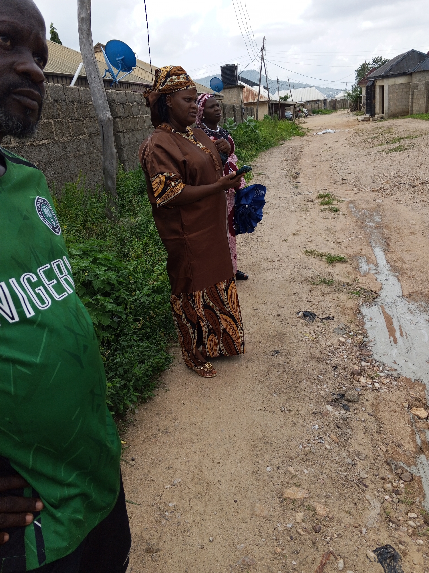 In Zango-ndaji, Nigeria, a man wearing a green shirt with white text that reads "NIGERIA" stands on the left side of a dusty dirt road. He is partially visible from the shoulders up, facing towards the right. To his right, two women are standing. The woman in the foreground is wearing a brown traditional dress with an ornate patterned hem and a headscarf of the same material. She is holding a dark blue umbrella in her right hand and a smartphone in her left. The second woman, behind the first, is wearing a pink and white patterned dress and a white headscarf. They are positioned on the side of the road where a shallow puddle of muddy water is present. The background reveals a settlement with various buildings and some satellite dishes. The sky is overcast.
