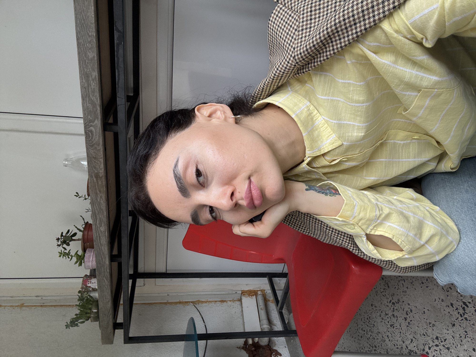 The image depicts a woman with dark hair and fair skin, wearing a yellow striped shirt and a brown plaid jacket. She is sitting on a red chair indoors, resting her head on her hand. A black metal shelf with plants is visible in the background. The floor appears to be a concrete or terrazzo surface. The location is most likely indoors in Tbilisi, Georgia, based on the architectural style and the context of the image.