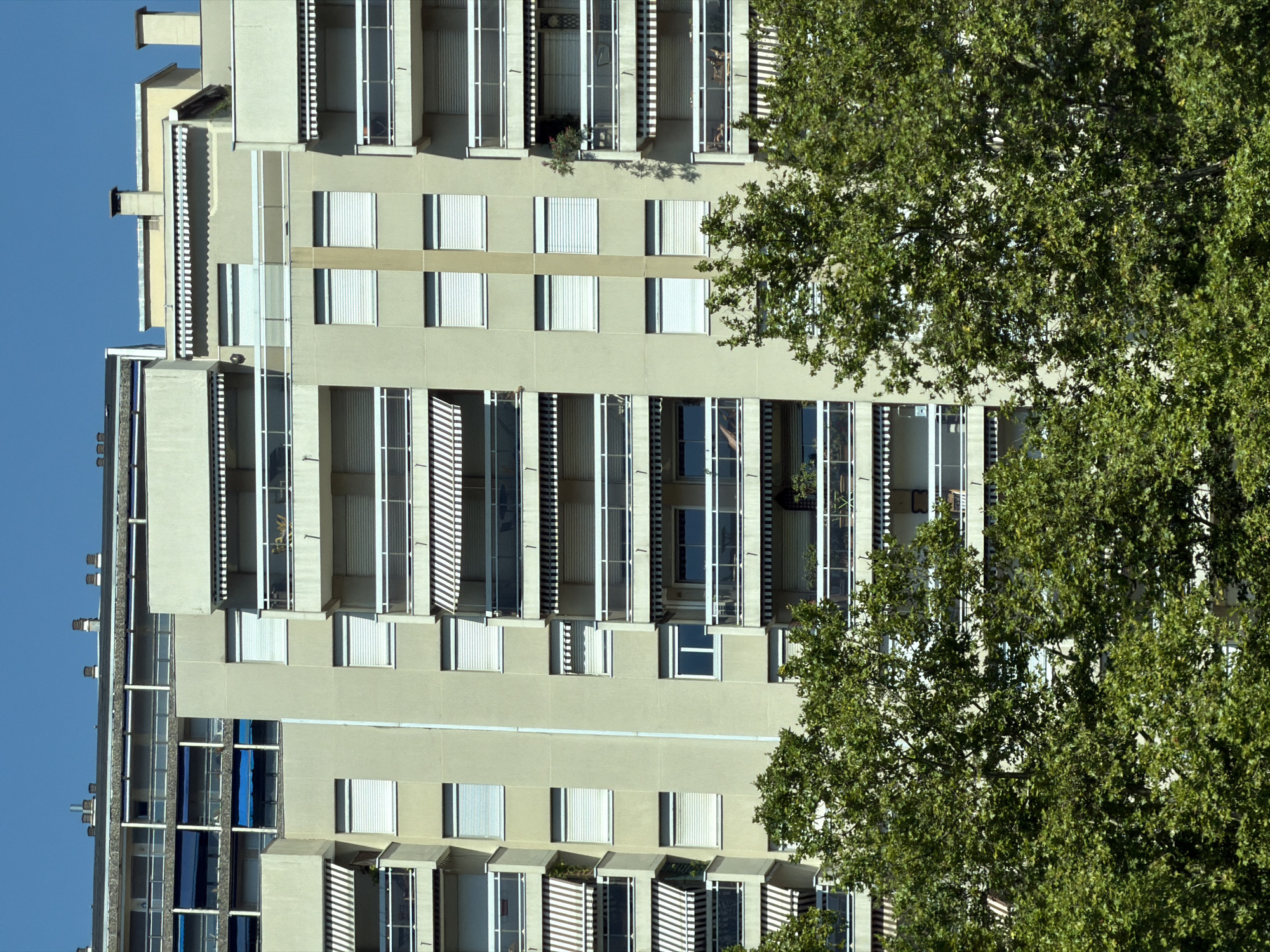 The image captures a portion of a large, multi-story apartment building in Lyon, France, set against a clear blue sky. The building is constructed with a light-colored facade and features numerous balconies with white railings. Many of the balconies are equipped with striped awnings in black and white, or solid white. The windows are predominantly covered by white roller shutters. Lush green trees with dense foliage partially obscure the lower levels of the building. The architecture suggests a mid-to-late 20th-century design, characteristic of many urban residential buildings. There are no people or visible activities depicted in the scene. The overall impression is one of a typical residential urban landscape.