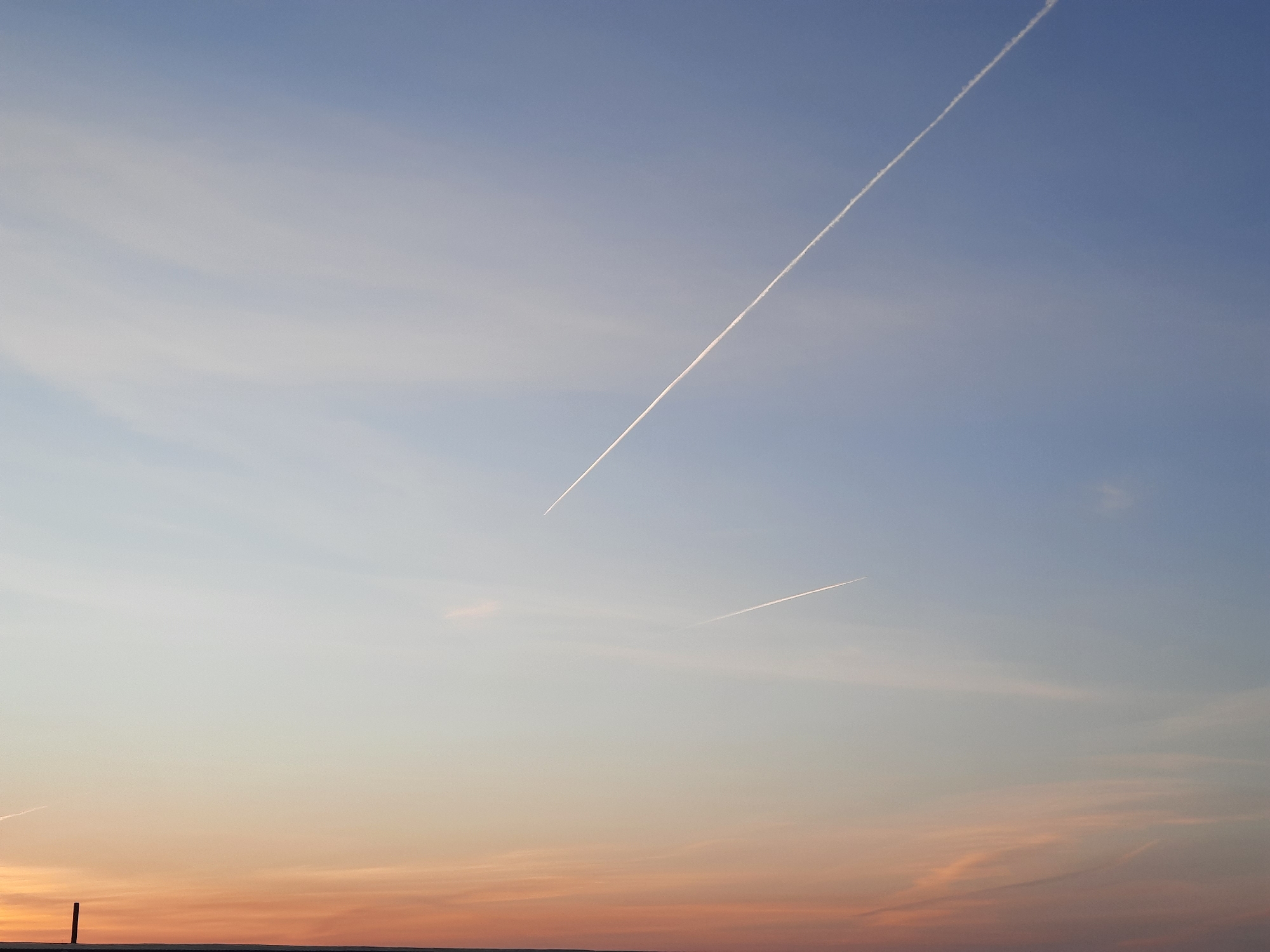 The image shows a sky at dusk or dawn. The sky transitions from a light blue at the top to orange and pink near the horizon. There are two prominent contrails from aircraft visible, one longer and higher in the frame, and the other shorter and lower. The horizon shows a faint strip of land. A thin vertical structure, likely a pole, is faintly visible at the far left edge of the frame. The scene is located in the city of Krasnogorskoye, Russia.