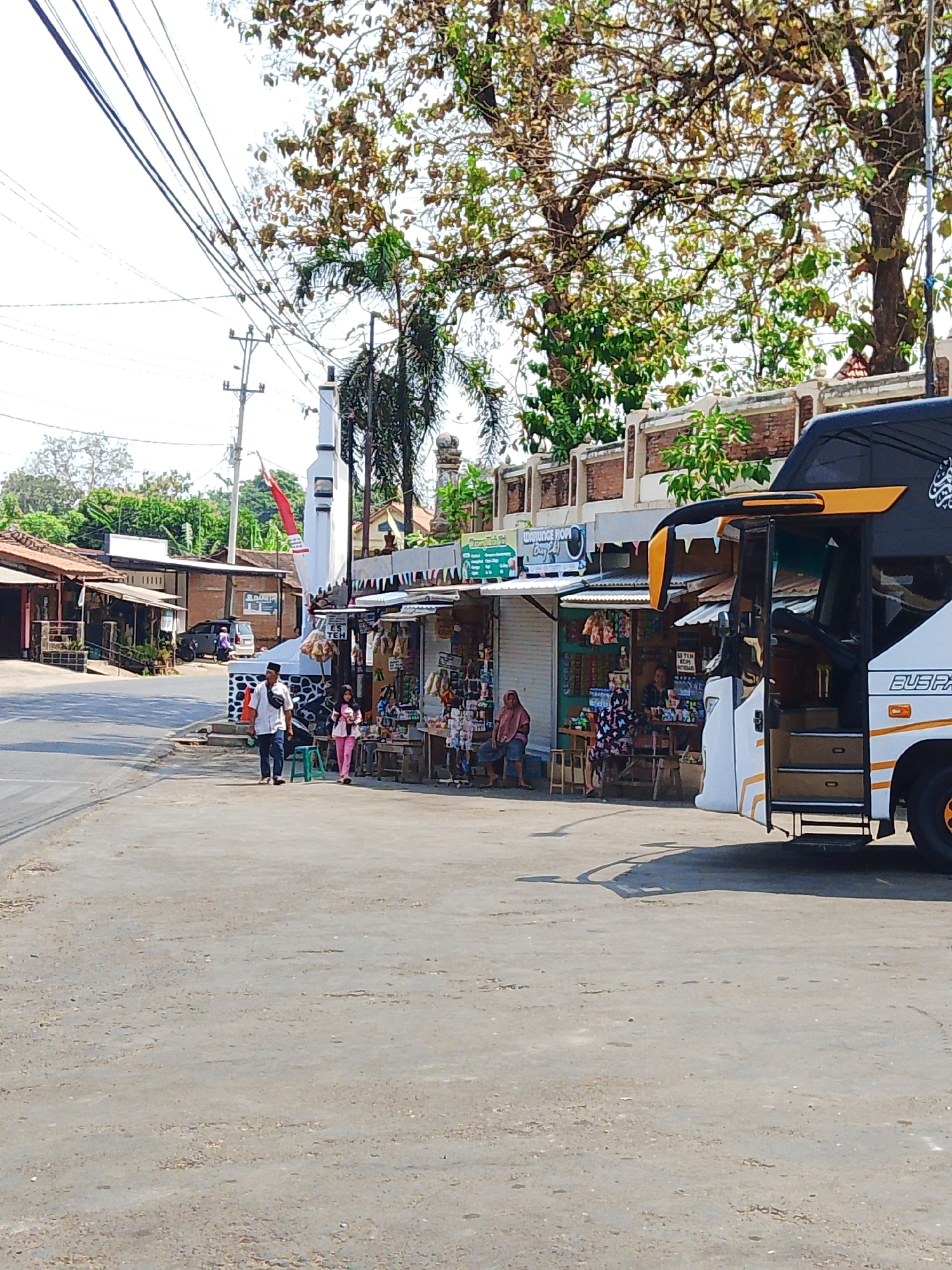 The image depicts a street scene in Mantingan, Indonesia, featuring a roadside market with several stalls. A white and black bus with orange accents is parked on the right side of the image, its door open. Two individuals, a man in a white shirt and dark pants and a woman in pink attire, walk towards the viewer on the paved road. A white monument with a red and white flag is located in the background. The market stalls display various goods, and several people are seen browsing or tending to the shops. The environment appears to be sunny with trees and overhead power lines contributing to the outdoor setting.