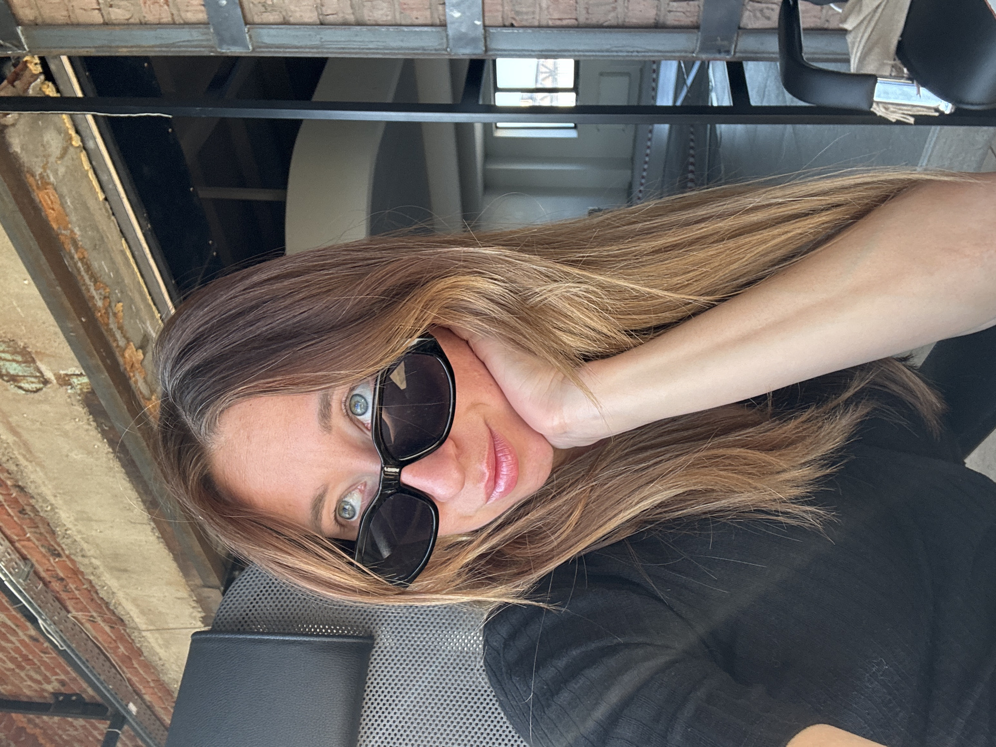 A fair-skinned woman with light brown hair and wearing sunglasses poses for a selfie. She is wearing a black top. The location appears to be indoors, with a view of a building and the sky through a window. Construction elements are visible, including brick walls and metal beams. The location within the city of Chaikovsky, Russia, is not evident.