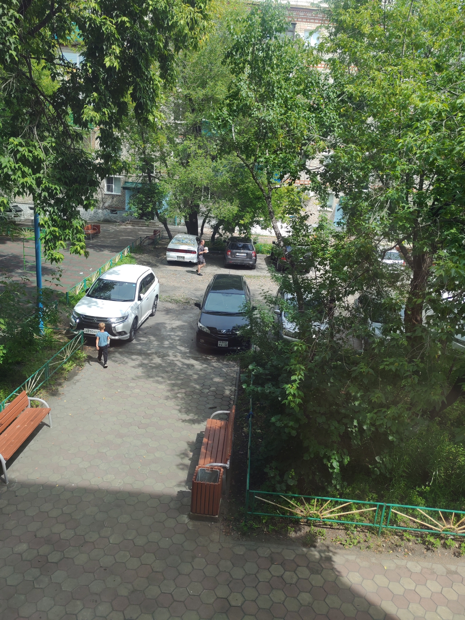 The image captures a scene in a residential courtyard in Belogorsk, Russia, on a sunny day. The foreground features a hexagonal-tiled paved area with two wooden benches and a trash receptacle. A green metal fence with decorative sunburst patterns separates the paved area from a grassy area with dense foliage and trees. In the middle ground, a white SUV is parked, and a young boy in a blue shirt and dark pants is walking away from it towards the paved area. Several other cars, including a black sedan and a white sedan, are parked further back in the courtyard. Trees with lush green leaves dominate the background, partially obscuring the multi-story brick buildings that appear to be apartment complexes. The overall impression is of a typical urban residential setting.