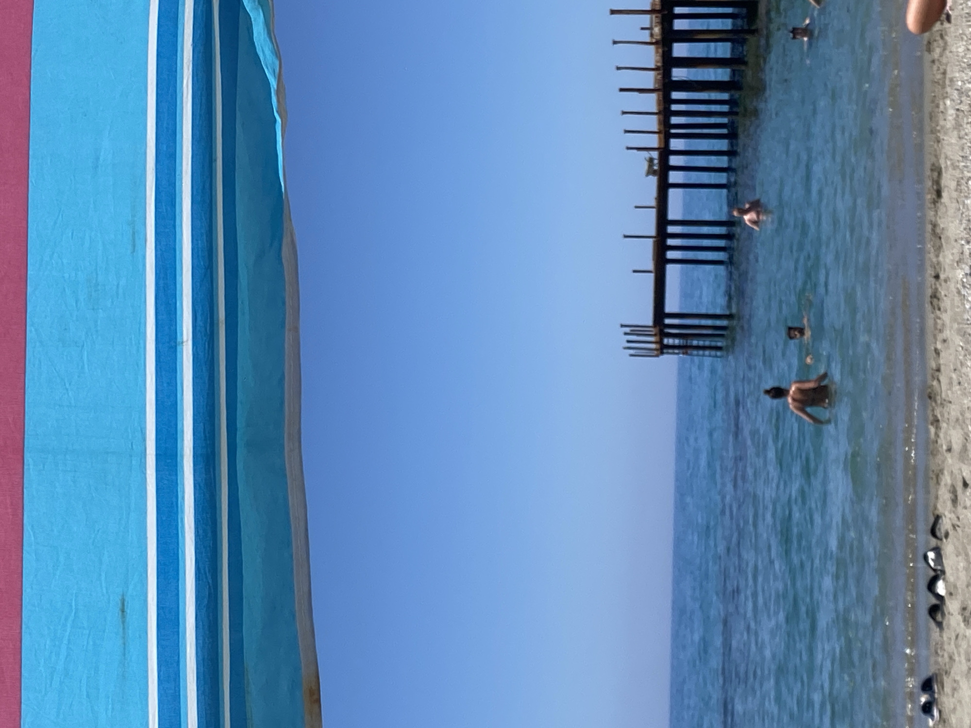 The image depicts a beach scene in Alanya, Turkey. A portion of a beach umbrella with blue, white, and red stripes is visible on the left. In the center is a clear blue sky. To the right, the turquoise sea meets a light-colored, pebbled beach. An old, decaying pier extends into the sea with visible rusted metal supports. Several people are swimming in the water near the pier. Footwear is scattered on the beach near the shoreline.