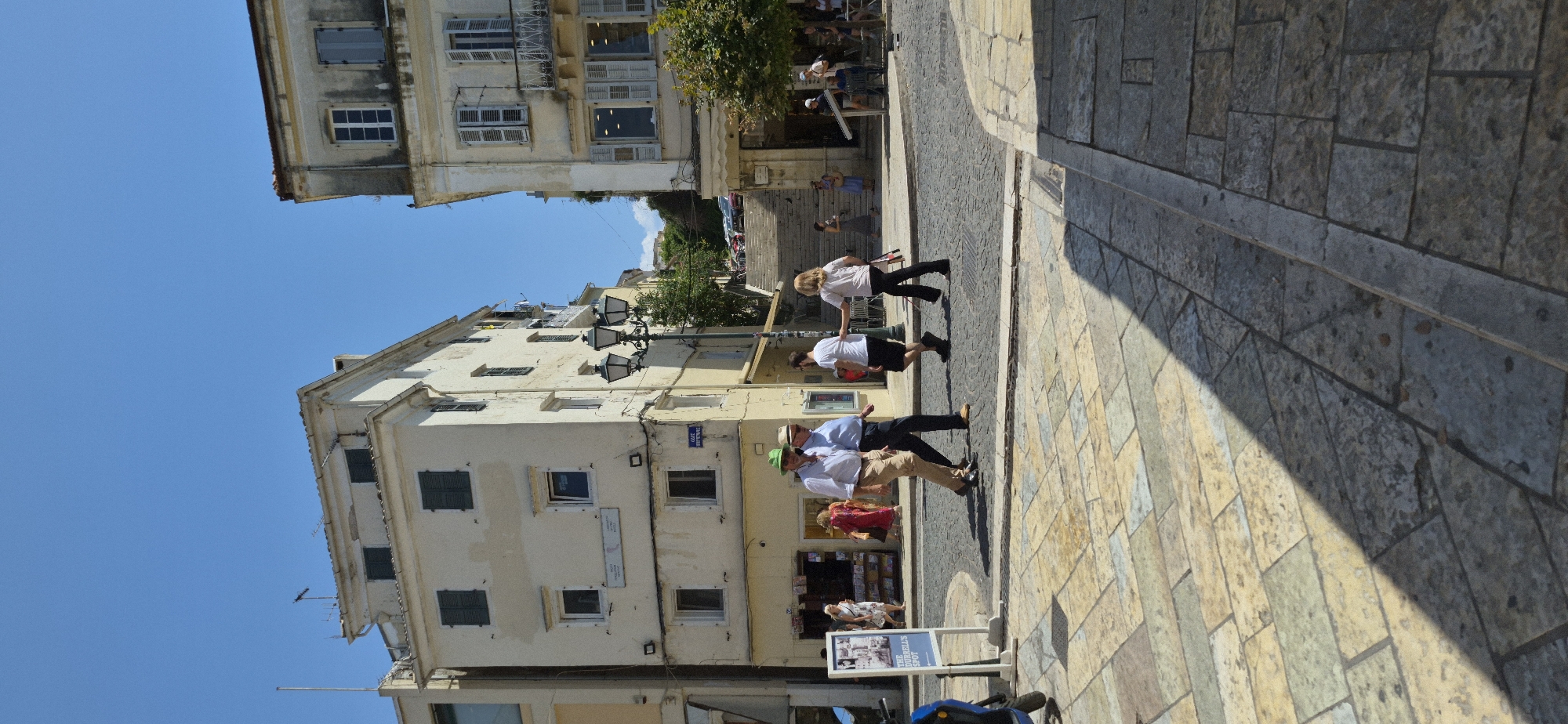 The image depicts a street scene in the city of Corfu, Greece, likely on a sunny day. In the foreground, a cobblestone street with a patterned paving in sections leads the eye towards the center of the frame. A shadow from an unseen object falls diagonally across the paving.

In the mid-ground, several people are walking. Two men in the center are walking together; one is wearing a white shirt, light brown pants, and a green hat, while the other wears a light blue shirt and dark pants. To their right, a woman in a white top and dark pants walks, and to her left, a man in a white t-shirt and black shorts walks in the opposite direction. Further back, another woman in a light-colored dress is visible, as well as other individuals in the distance.

The background features buildings with traditional architecture. To the left is a multi-story building with cream-colored walls and dark shutters. To the right, another building has a similar color scheme, with balconies and many windows. A set of stone stairs is prominent in the background, leading upwards, with some vehicles visible at the top. Street lamps are also visible, adding to the urban setting. A sign with images and text, likely an advertisement, is positioned on the left side of the image. The sky is a clear blue.