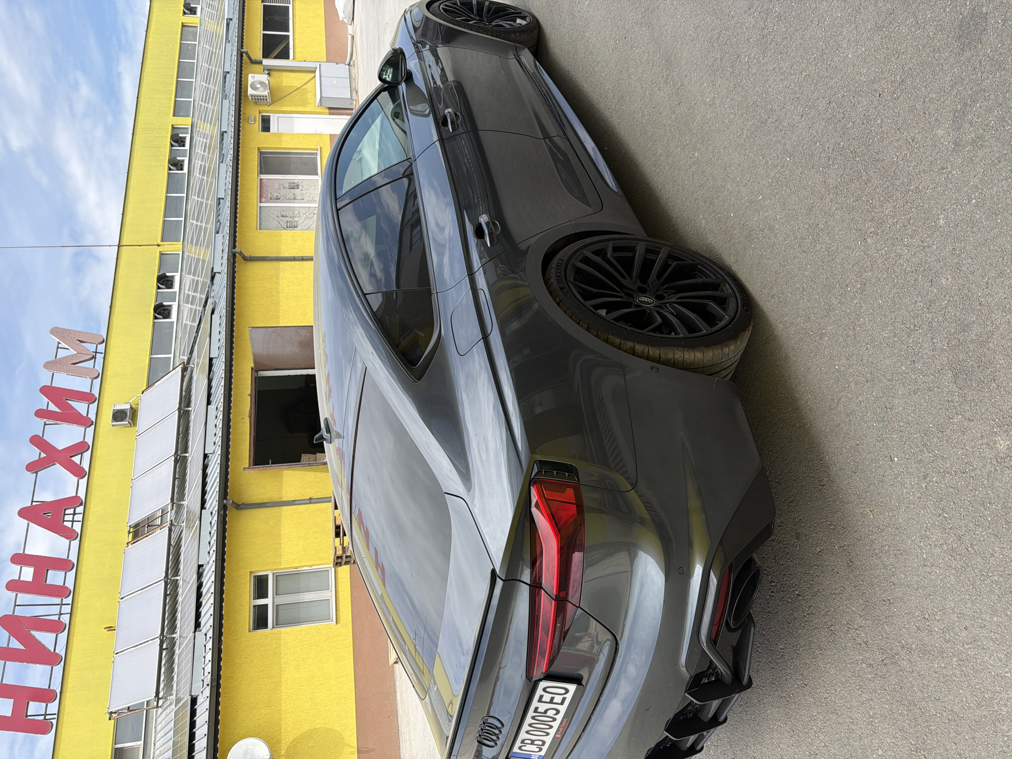 The image depicts a gray Audi parked on an asphalt surface next to a yellow building with a sign in Cyrillic. The building features numerous windows, some with blinds. An air conditioning unit is visible on the building's facade. The car is angled in the frame, showcasing its rear and side profile. The license plate number of the car is visible and reads "CB 0005 EO". The overall setting suggests an urban environment, possibly in Dolno Ablanovo, Bulgaria.