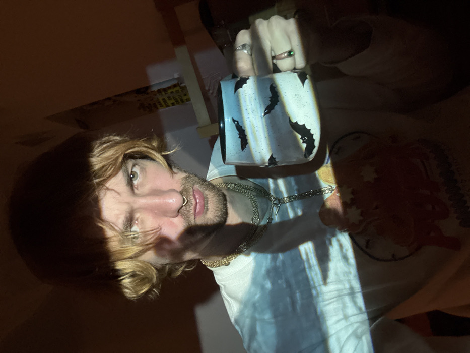A person with shoulder-length blonde hair and a nose ring holds a mug with bat designs. The person wears a white t-shirt and two layered necklaces. A hand with rings is visible holding the mug. The scene is dimly lit with strong contrast, creating patches of light and shadow across the person's face and clothing. Part of a blanket with a circular design is visible.