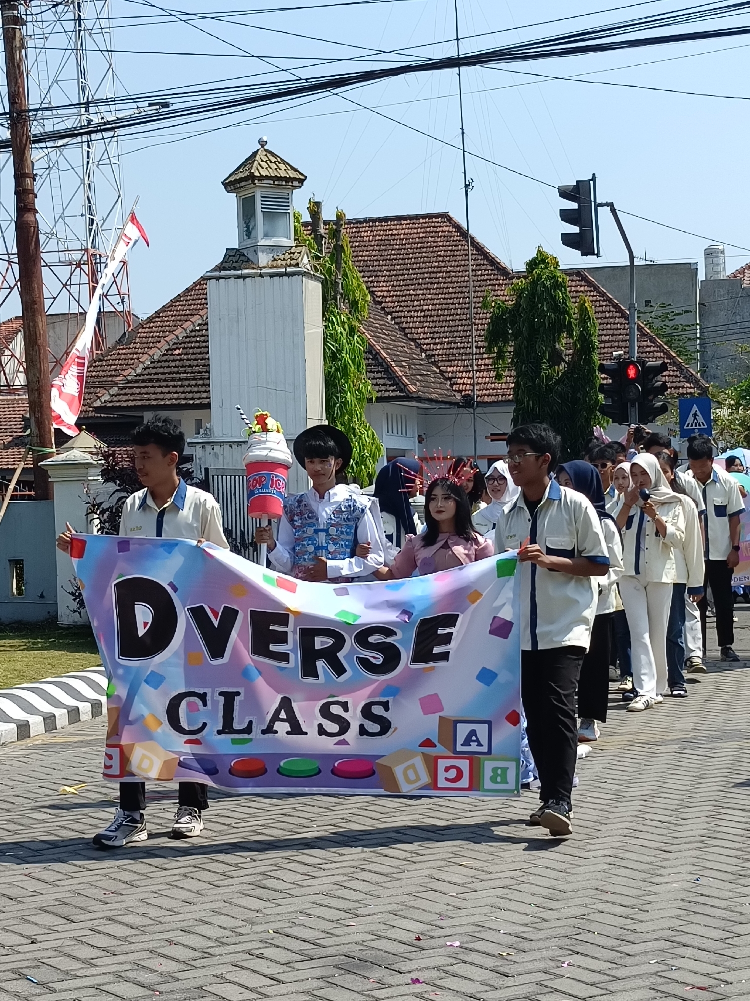 The image depicts a parade scene in Kediri City, Indonesia. A group of young people, likely students, are walking in procession, holding a banner that reads "DIVERSE CLASS." The banner is colorful and adorned with confetti and building blocks. Two individuals in the foreground are prominently featured, one on the left holding the banner, and another on the right carrying a large, cylindrical object with a depiction of an ice cream cup and a topping.
The participants are dressed in various attires, with some wearing uniforms that appear to be school or organizational attire consisting of light-colored shirts and dark pants. Others are in more festive or traditional clothing. In the background, a street is visible with a brick-like pavement. Buildings with tiled roofs, utility poles, and power lines are also part of the urban environment. A traffic light is visible in the distance, indicating a road intersection. The sky is clear and blue, suggesting a sunny day. The overall atmosphere suggests a community event or celebration.