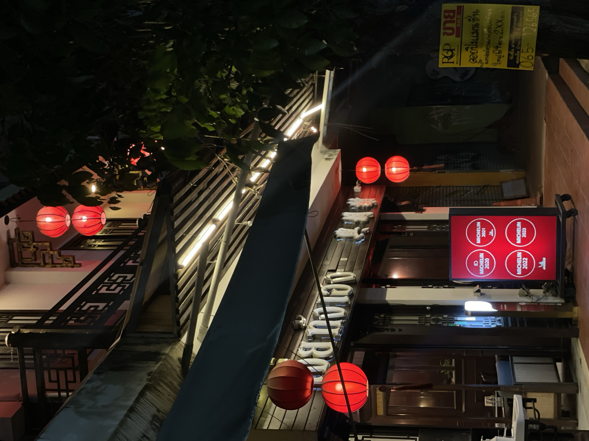 Captured at night in Chiang Mai City Municipality, Thailand, the image presents a street scene with an oriental theme. Red Chinese lanterns cast a warm glow, illuminating the exterior of what appears to be a restaurant or shop. A sign prominently displays "MICHELIN" with various years (2020, 2021, 2022, 2023) suggesting the establishment has received accolades. A sign advertising "ROP ขาย" (presumably sales or renting) in Thai, along with a phone number, is visible. Trees overhang the scene, adding depth and suggesting the building is nestled amongst foliage. The architecture has a distinct Chinese influence, and the lighting creates an inviting atmosphere.