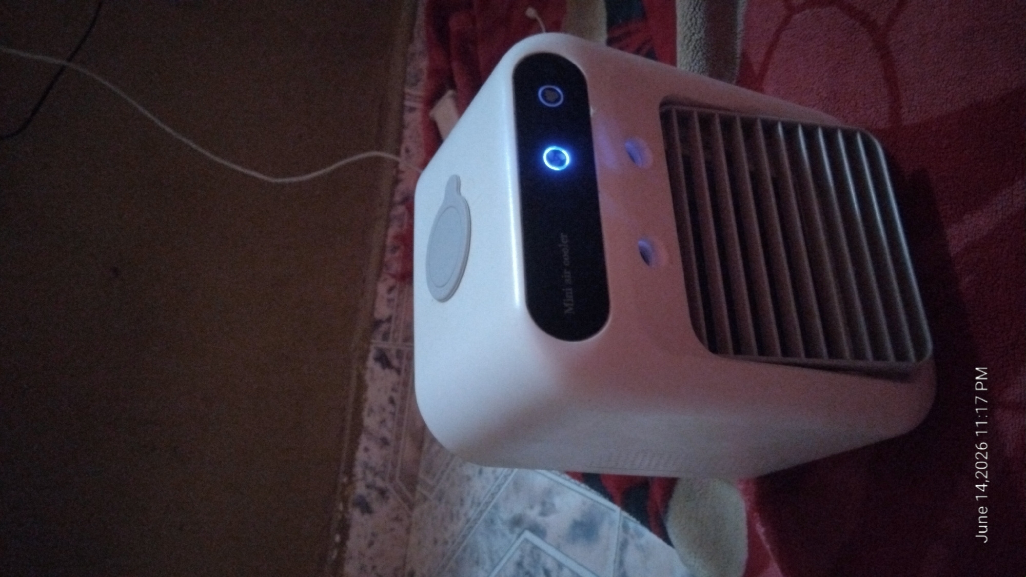 A white mini air cooler sits on a red patterned blanket. The air cooler has a gray lid on top and a black panel with "Mini air cooler" written on it. There are two small blue lights on the front of the air cooler, and a circular blue light is illuminated on the black panel. The background is dark and blurry, with a brown wall and some wires visible. A timestamp in the bottom left corner reads "June 14, 2026 11:17 PM". The scene appears to be indoors.