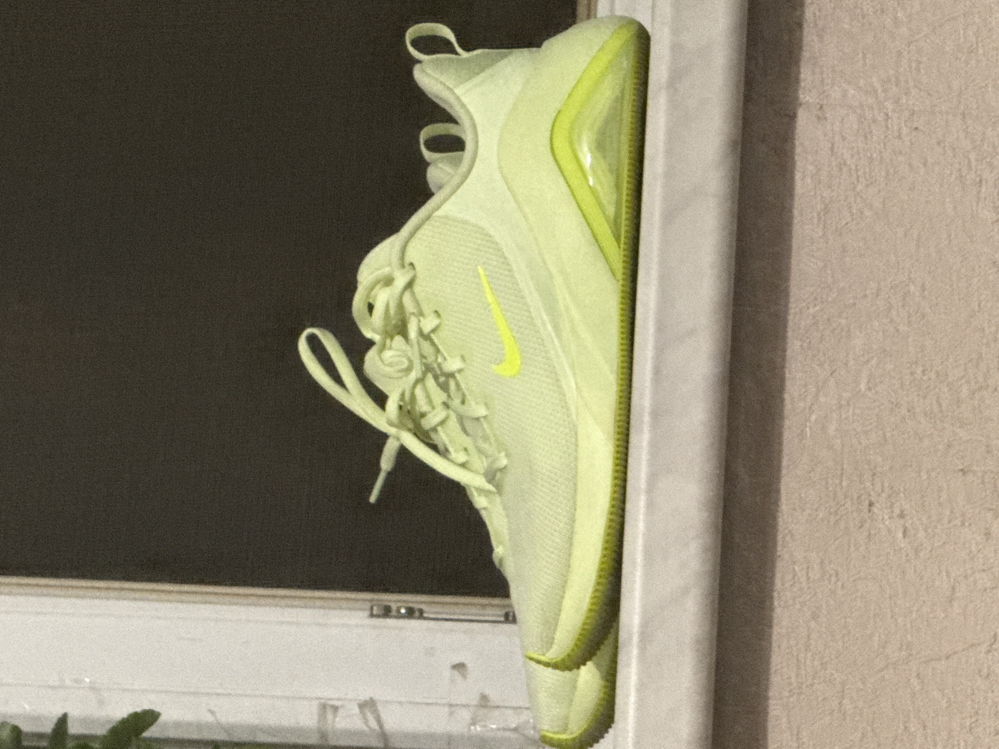 A pair of neon yellow Nike sneakers are placed side-by-side on a white ledge. The sneakers are oriented to the left, with their laces tied loosely. The background consists of a dark window and a white window frame. The ledge appears to be made of stone or concrete, with a textured wall visible below.