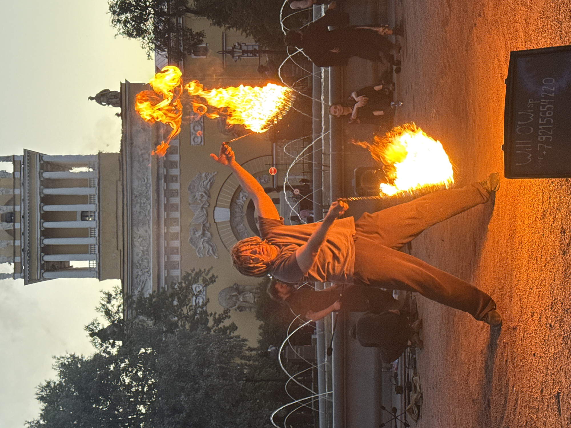 The image depicts a street performance in front of the Admiralty Building in Saint Petersburg, Russia, as indicated by the architectural style and the visible structure of the building. A fire performer, a person with dreadlocks wearing a grey t-shirt and grey sweatpants, is actively engaged in a fire-spinning act. They are holding a prop that is engulfed in flames, creating a large, bright fire that illuminates the surrounding area with an orange glow.
Several other individuals are present in the background. To the left of the performer, another person in dark clothing is partially visible, possibly assisting or observing. Further back, a fountain with multiple jets of water is active. In the mid-ground, to the right of the performer, a few spectators are seated, including a woman with dark hair wearing glasses, and another person in dark clothing. A person in a dark hooded cloak stands to the far right. The ground is covered with small gravel or sand, reflecting the fire's light. In the foreground, a dark rectangular sign with text written in white chalk is placed on the ground. The text appears to read "Will O'Wisp" and a phone number "+79215654220". The sky above is a pale yellow, suggesting either early morning or late evening. The overall scene captures a dynamic moment of urban entertainment amidst a historic architectural backdrop.
